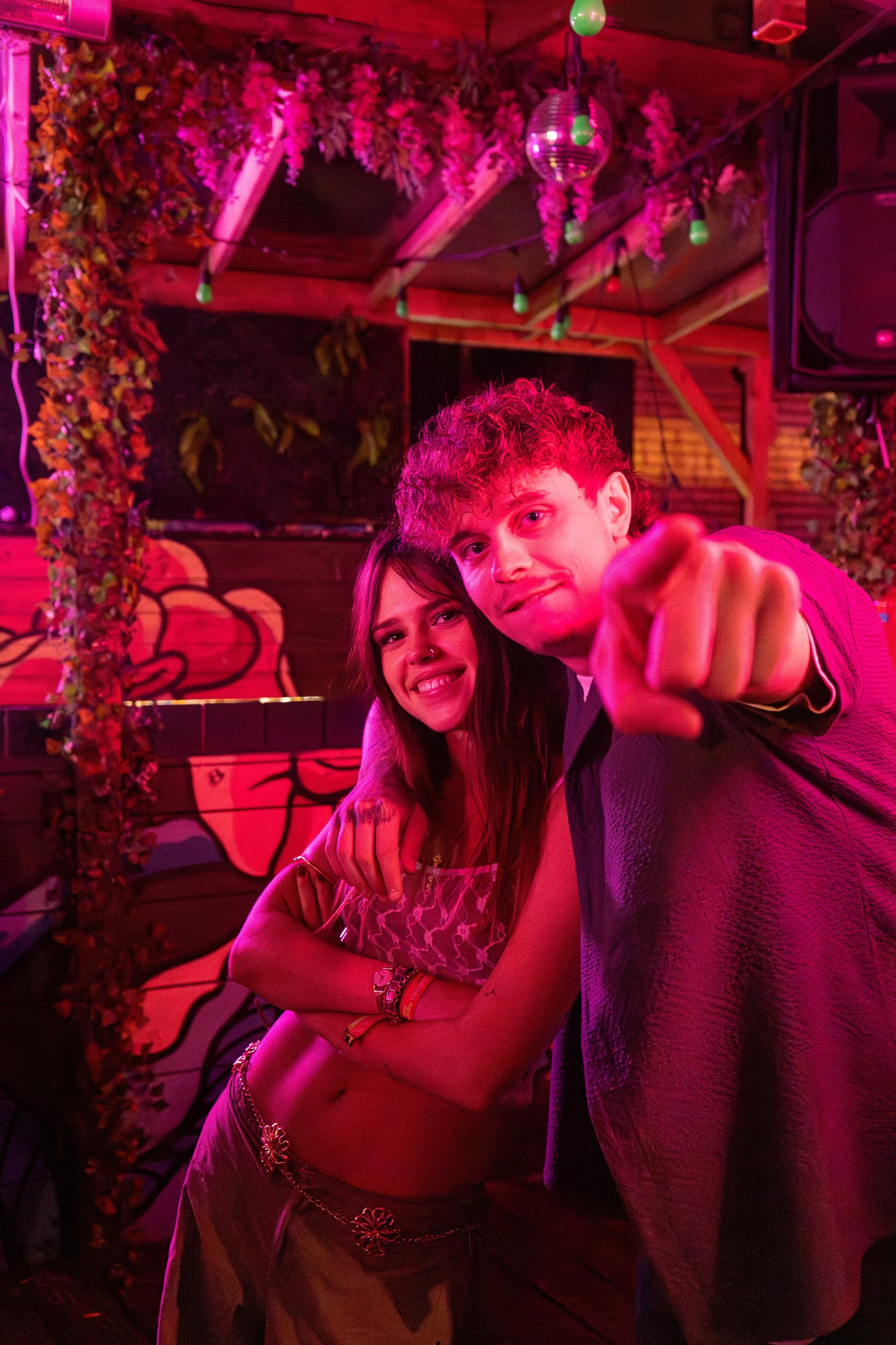 Two young people at a colorful, neon-lit party, with one pointing at the camera and the other smiling, in a decorated venue with plants, a mural, and a disco ball.