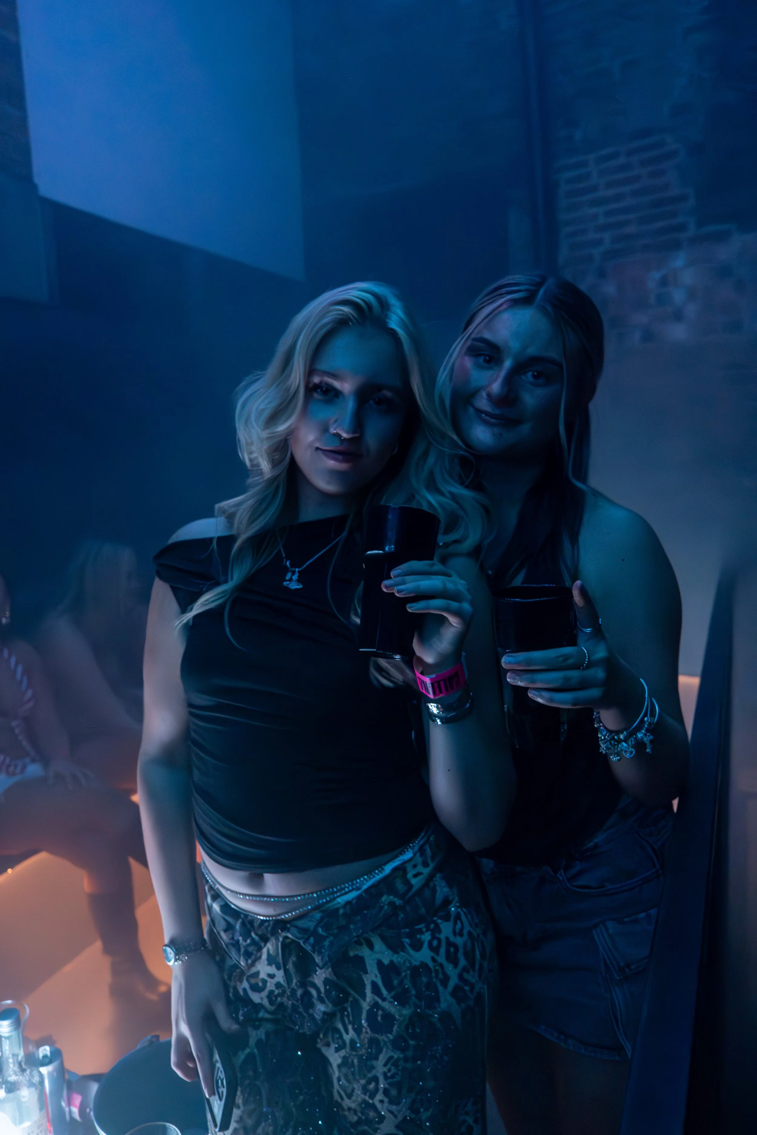 Two women at a party holding drinks and smiling, with blue lighting and a brick wall in the background.