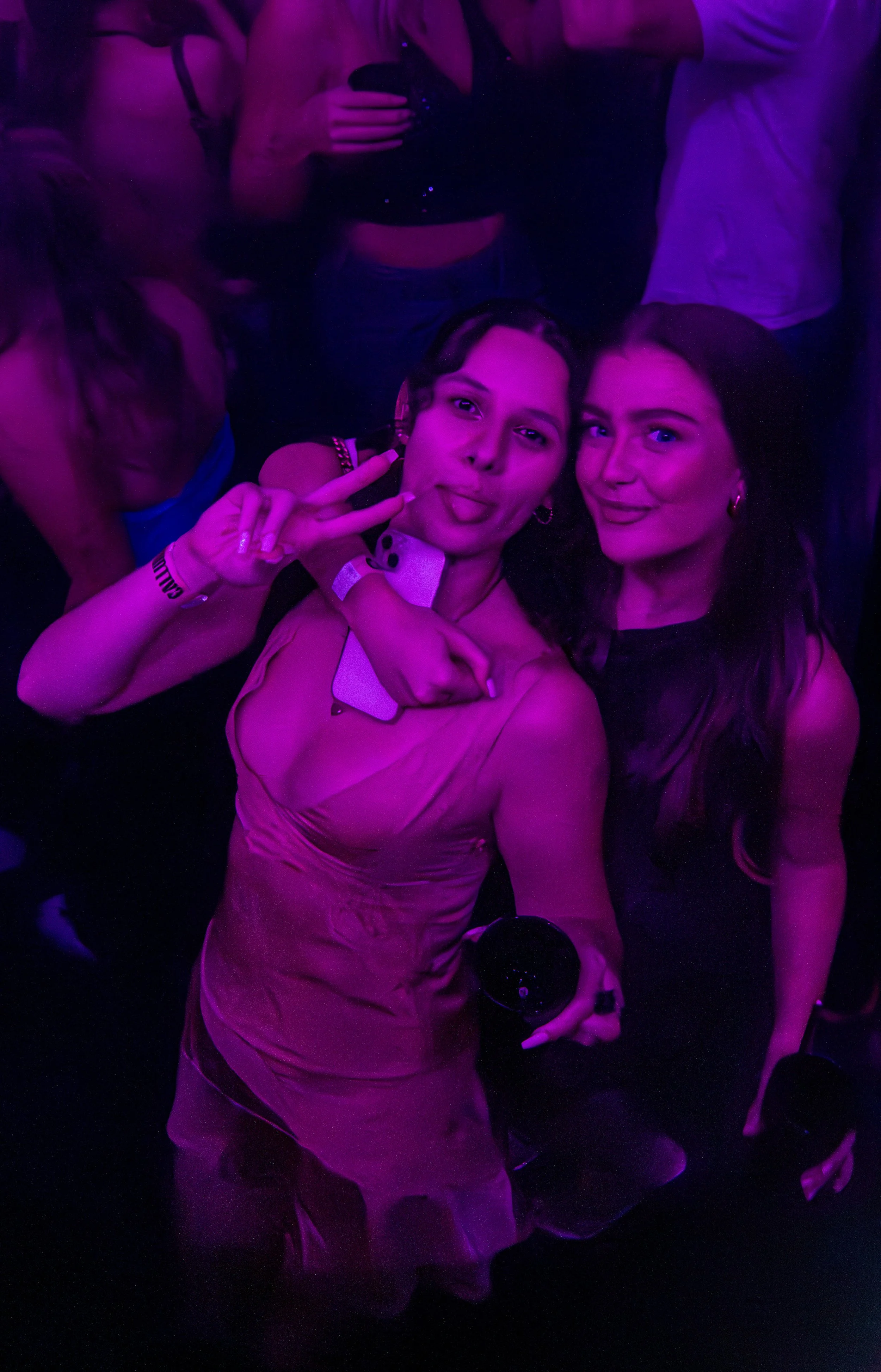 Two young women taking a selfie at a party with purple lighting, making peace signs and sticking out their tongues.