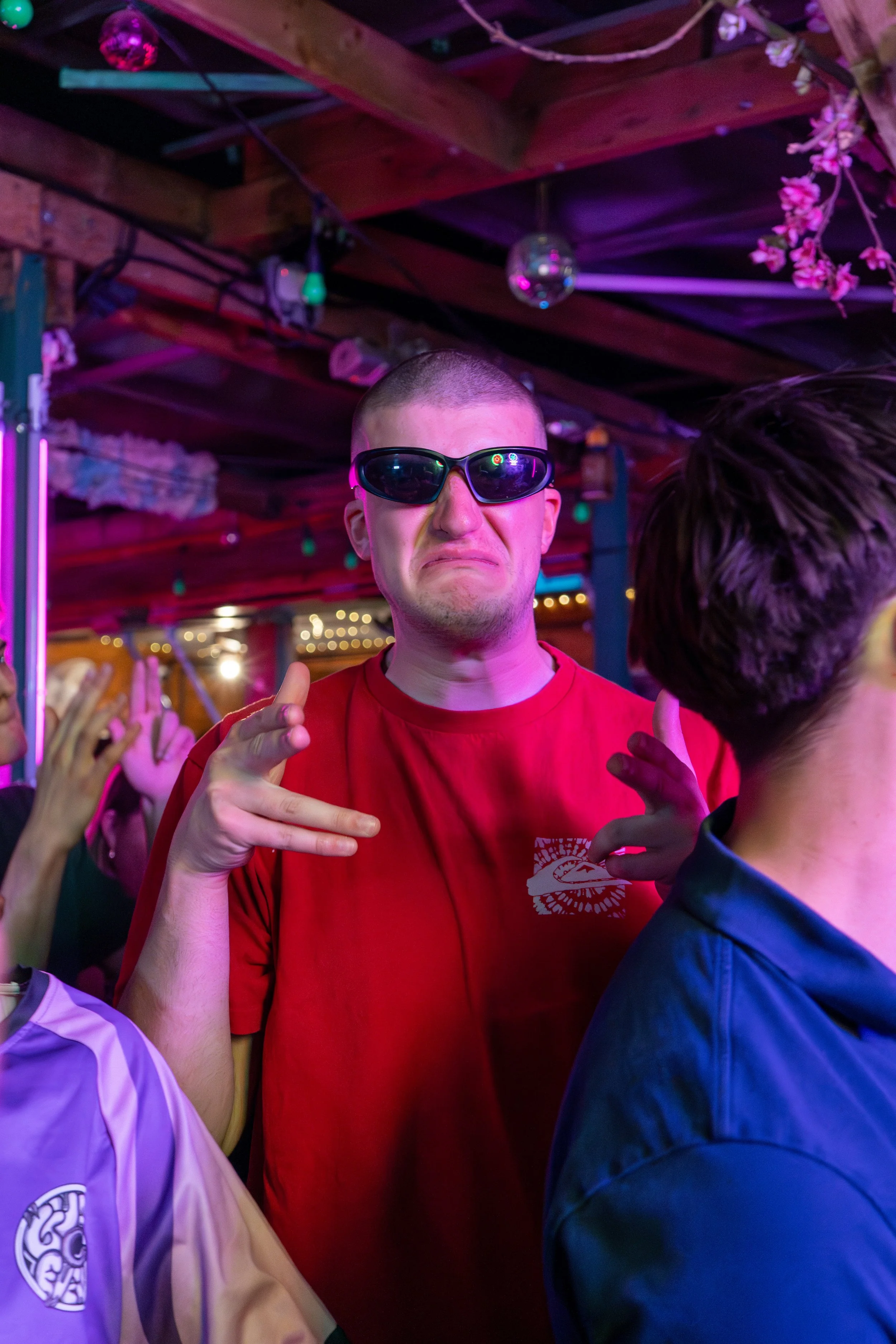 A young man wearing sunglasses and a red T-shirt at a party or nightclub, making a hand gesture, with colorful lights and other people in the background.