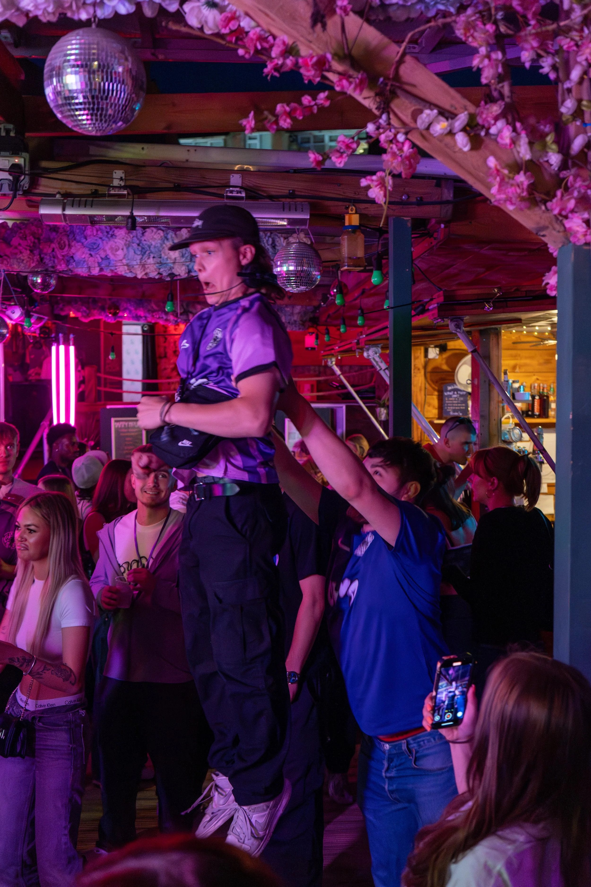 Group of people in a lively nightclub, with a person sitting on another person's shoulders, surrounded by colorful lights, floral decorations, and reflective disco balls.