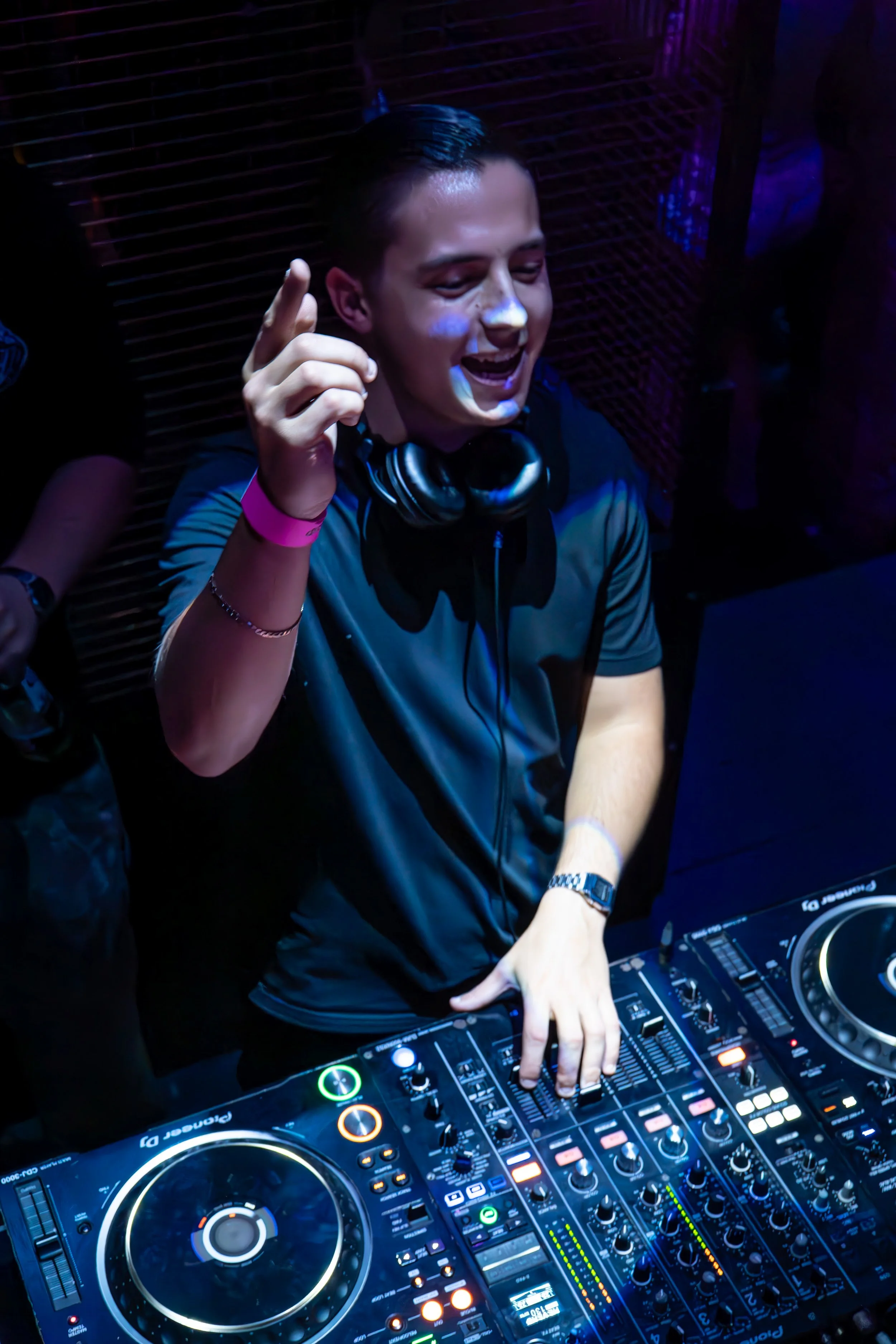 A male DJ with headphones around his neck, smiling and pointing upward, operating a DJ mixer under colorful club lighting.