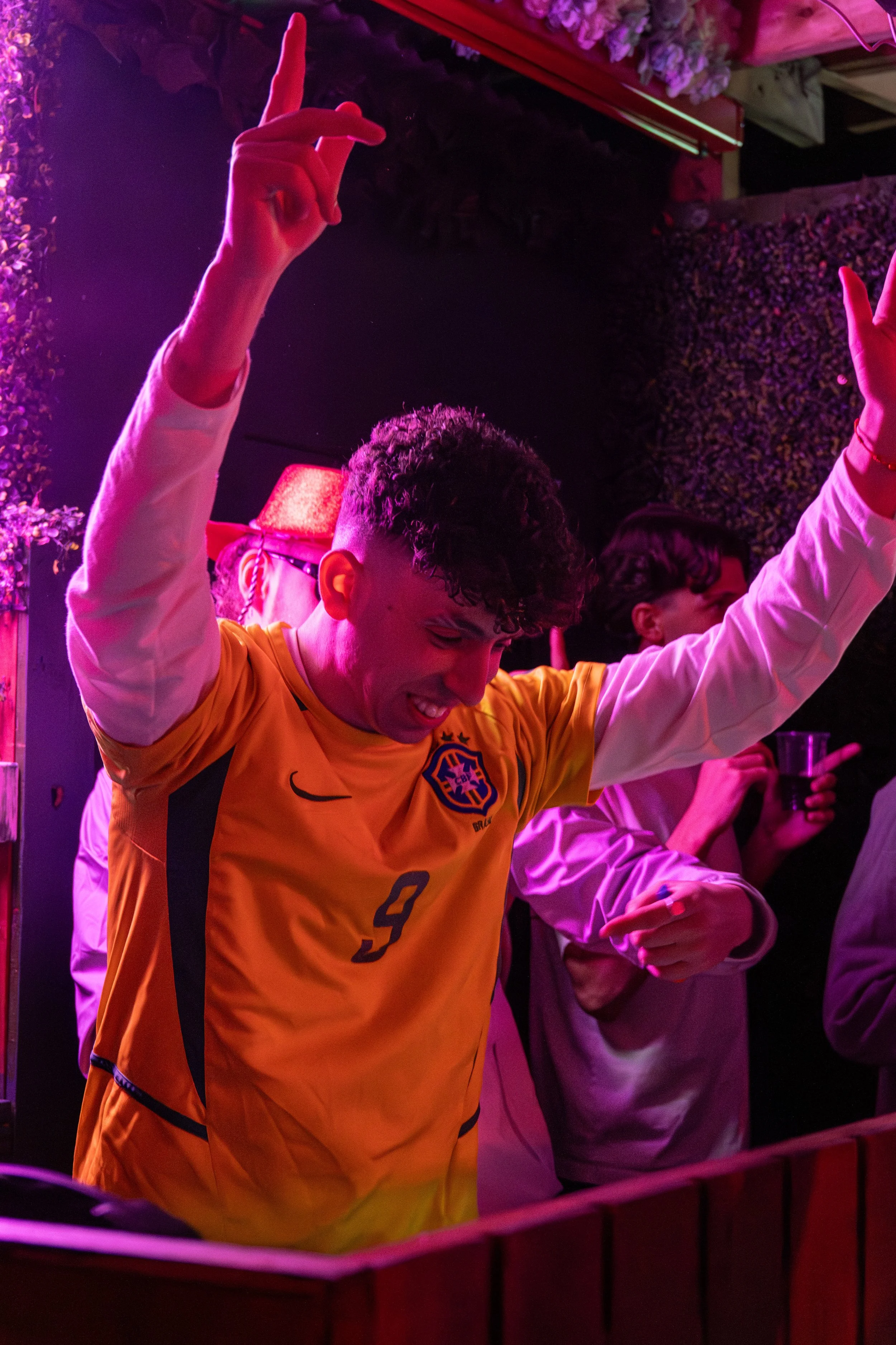 A young man wearing a yellow soccer jersey with the number 9, dancing with his arms raised and smiling in a lively party setting with pink/purple lighting.