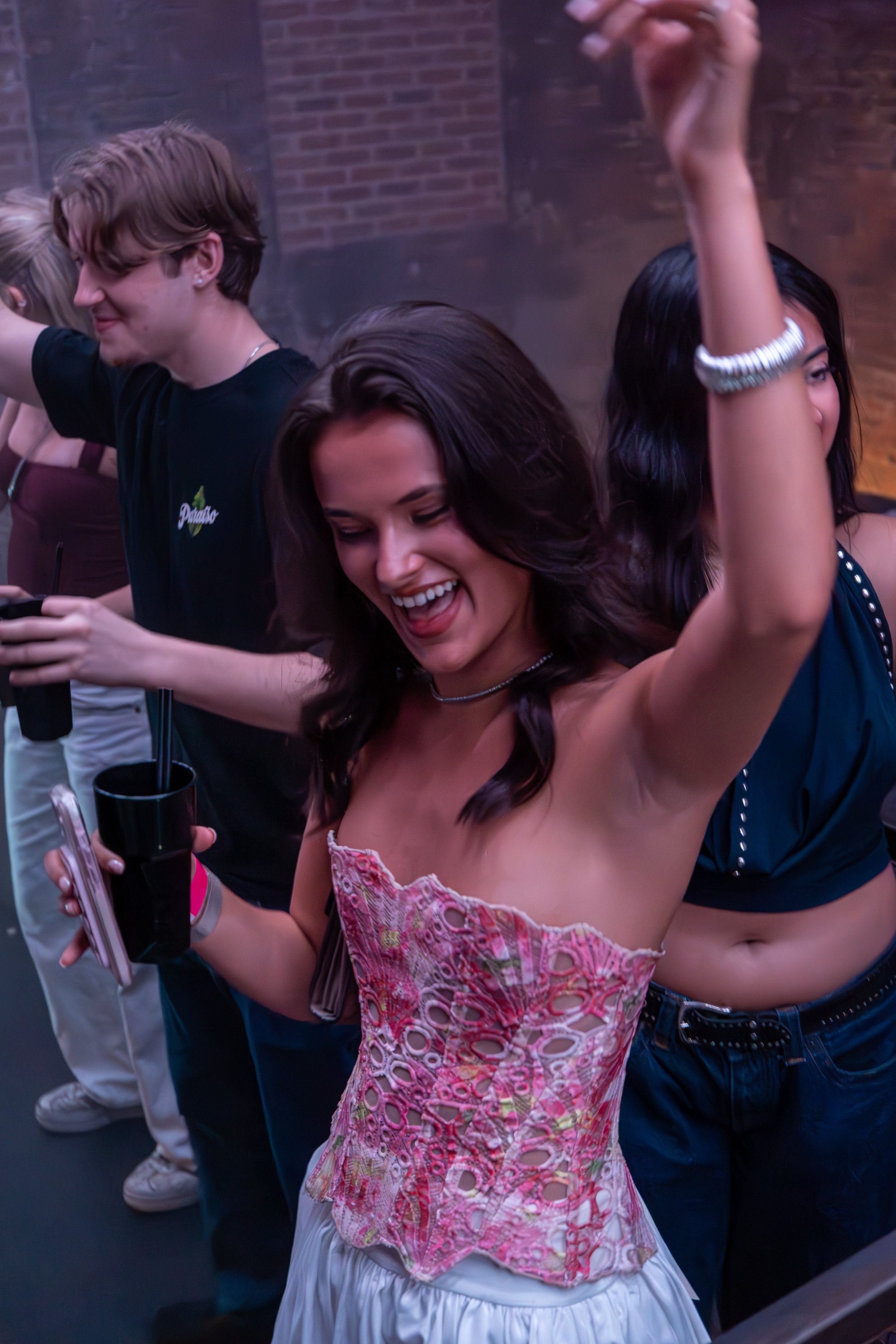 A woman in a pink and white patterned strapless top with her arm raised and a wide smile, enjoying a dance or party. Other people are in the background, also dancing or celebrating.