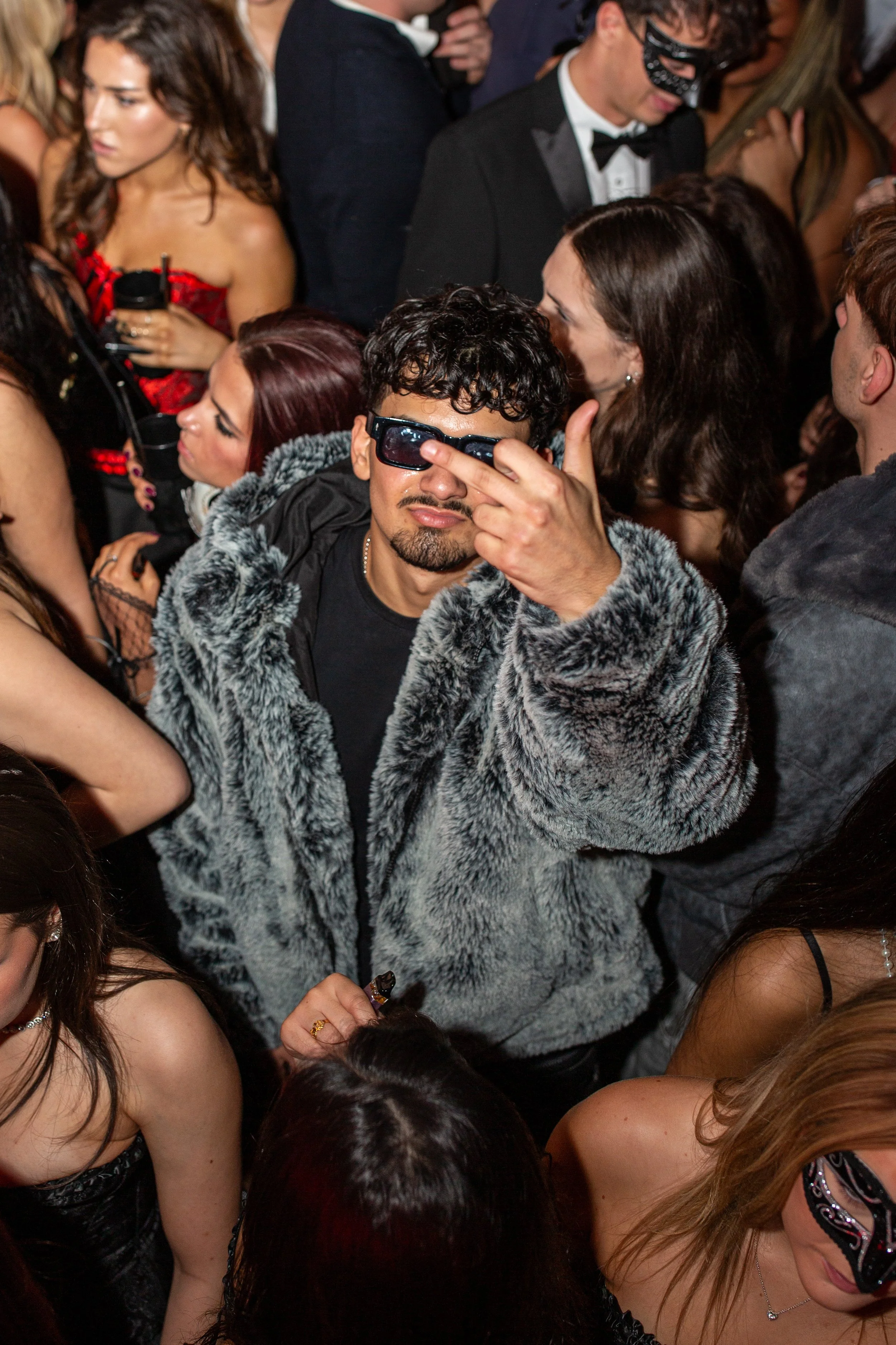 A man in a furry gray coat and black sunglasses making a gesture with his middle finger at a crowded party.