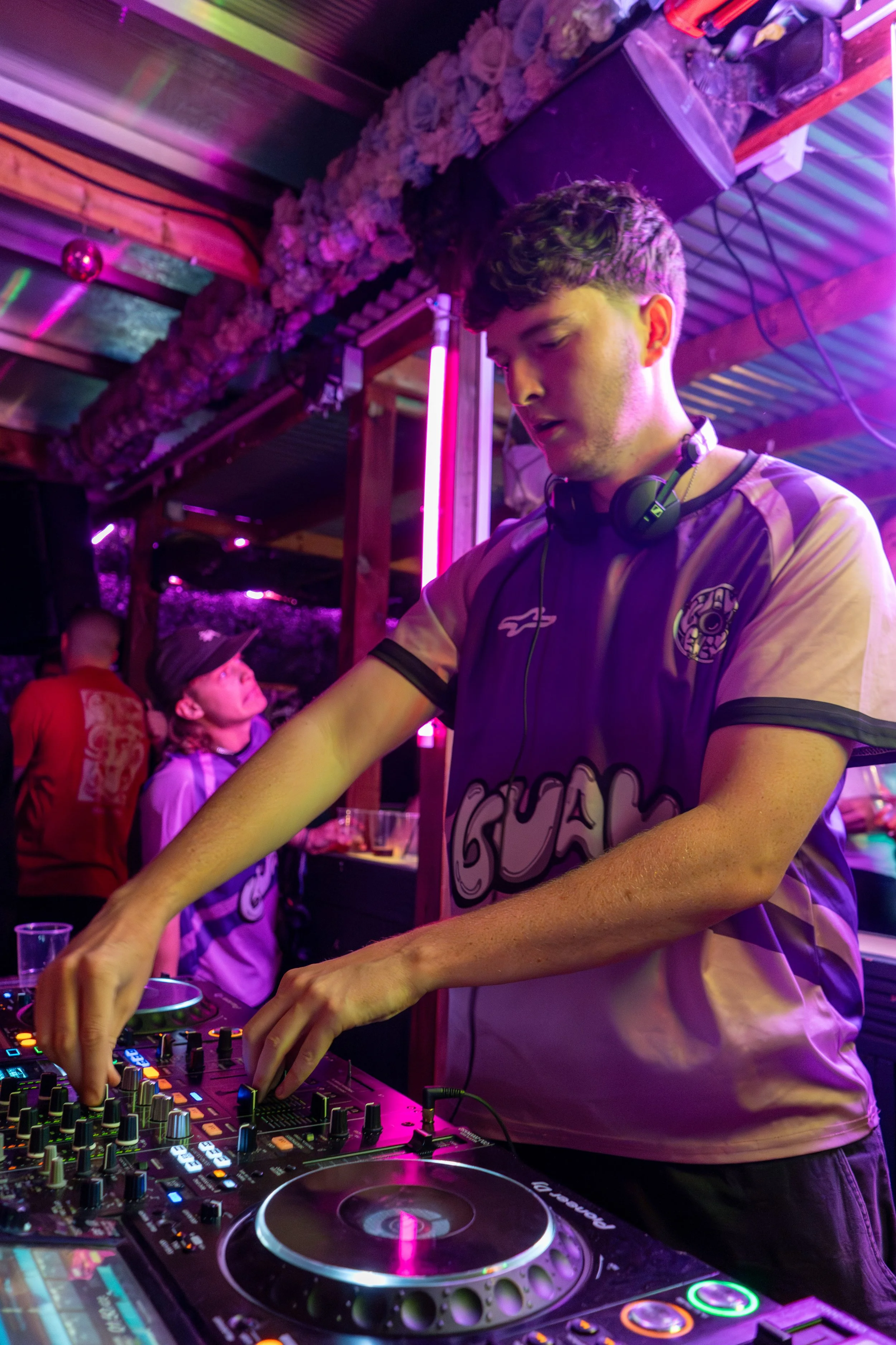 A young male DJ wearing a sports jersey with headphones around his neck, mixing music on DJ equipment in a colorful, vibrant club setting with pink and purple neon lighting.