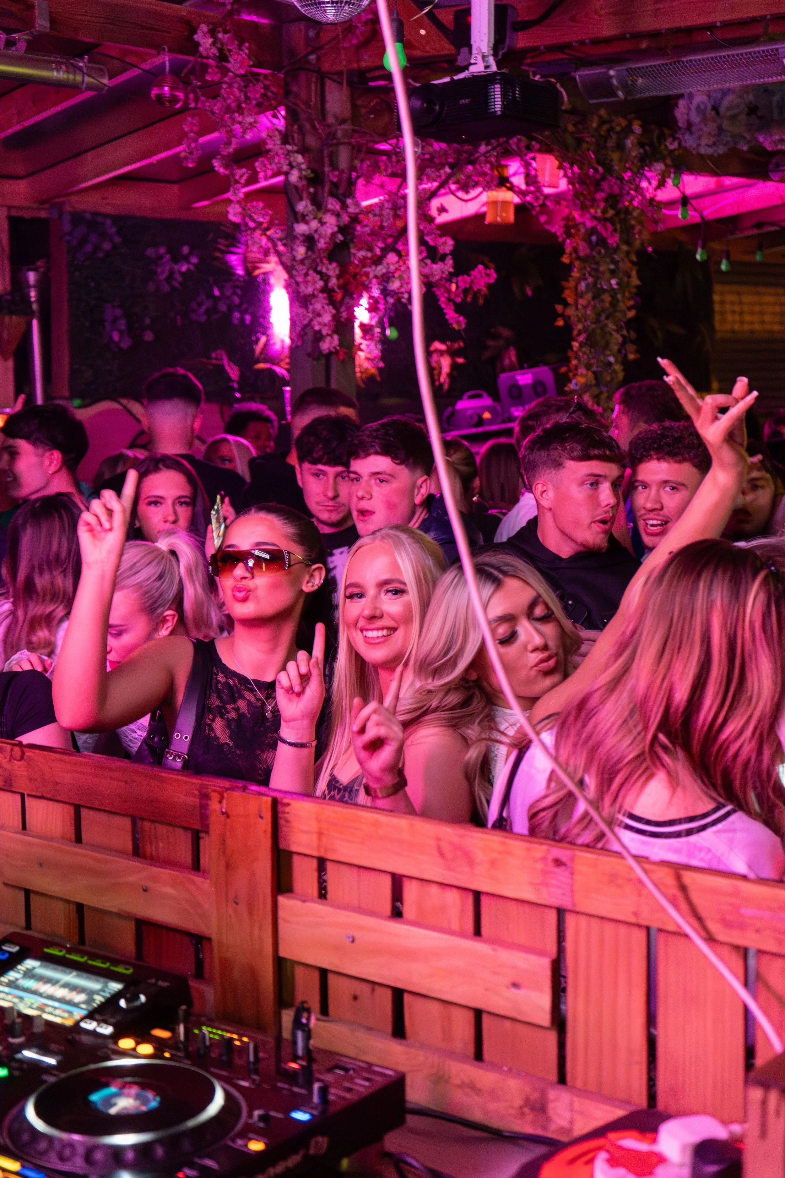 Young people dancing and enjoying a lively party scene with colorful lighting, DJ equipment, and decorative floral elements overhead.
