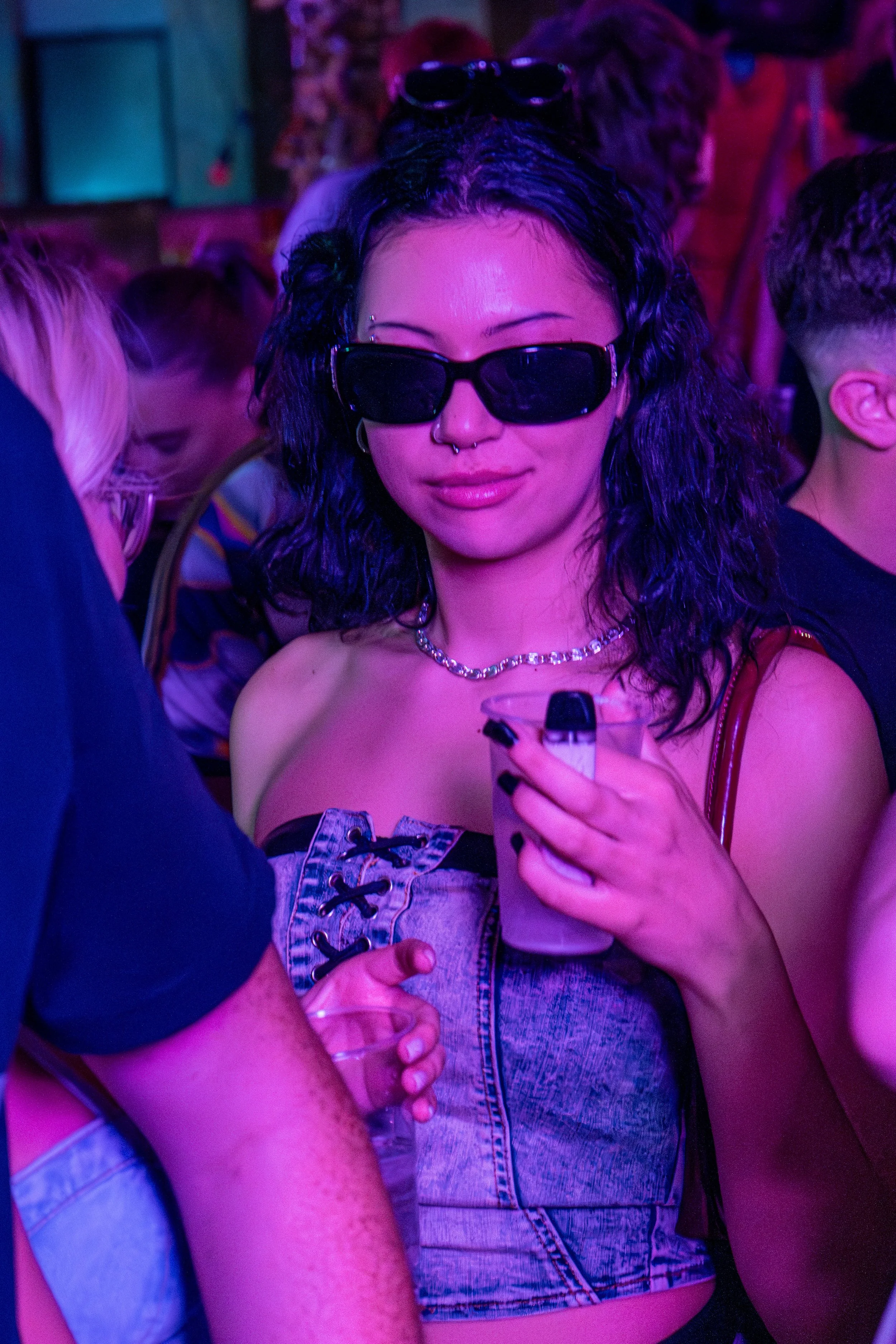 A woman with curly dark hair, wearing sunglasses, a silver chain necklace, and a denim sleeveless top, is holding a drink in a clear cup at a crowded party or club with purple and pink lighting.
