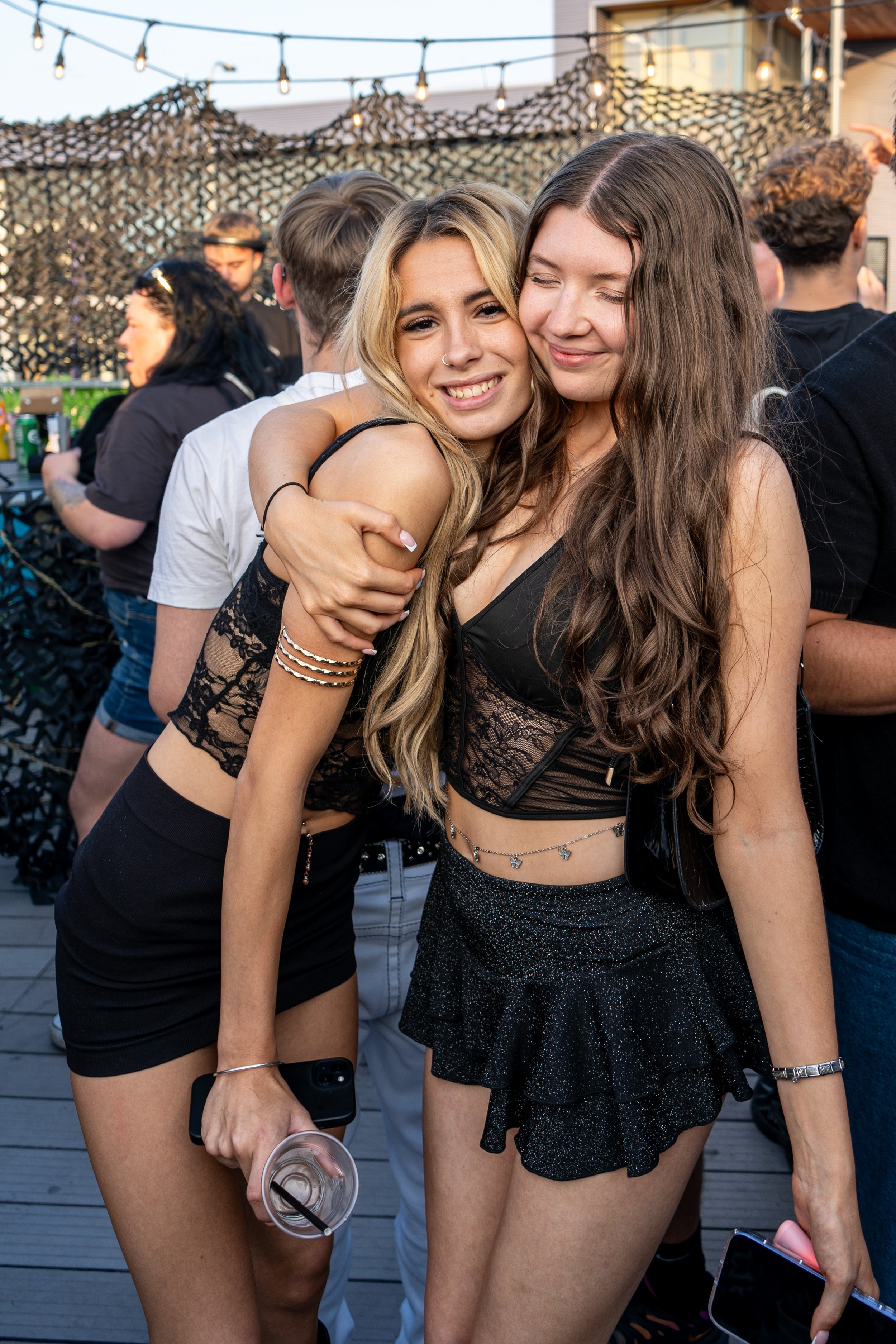 Two young women hugging and smiling at a social gathering on a rooftop with string lights and string netting overhead, surrounded by other people.