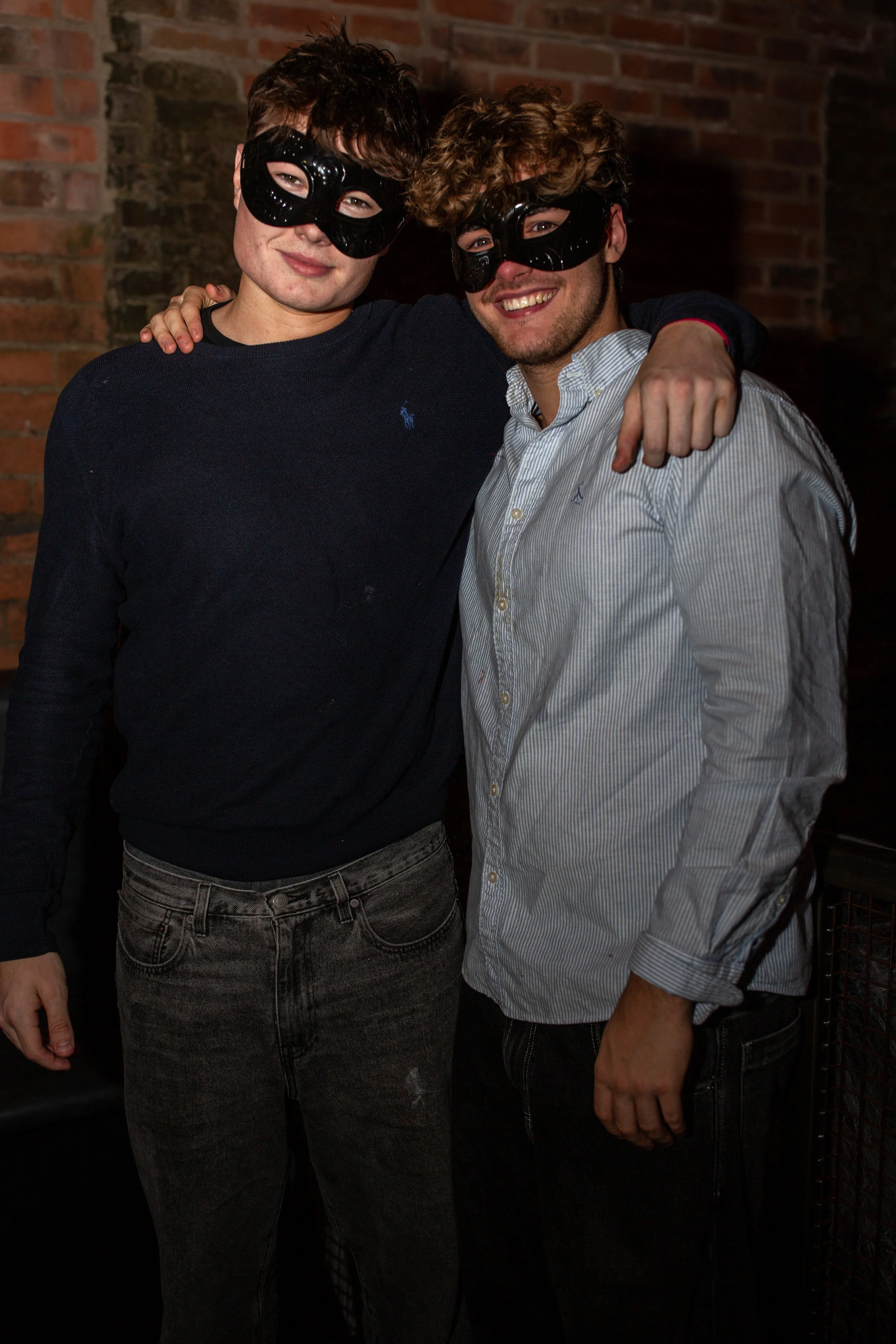 Two young men wearing black masks smiling and posing with their arms around each other in front of a brick wall.