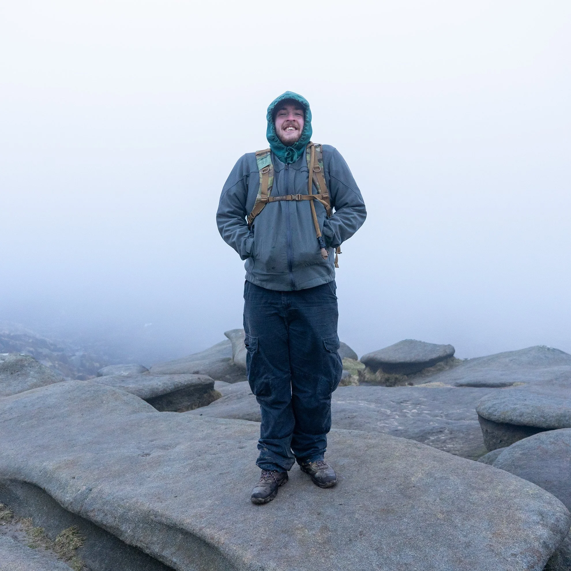 A man stands on a large rock surface outdoors in foggy weather, wearing a hooded jacket, cargo pants, hiking boots, and a backpack, smiling.