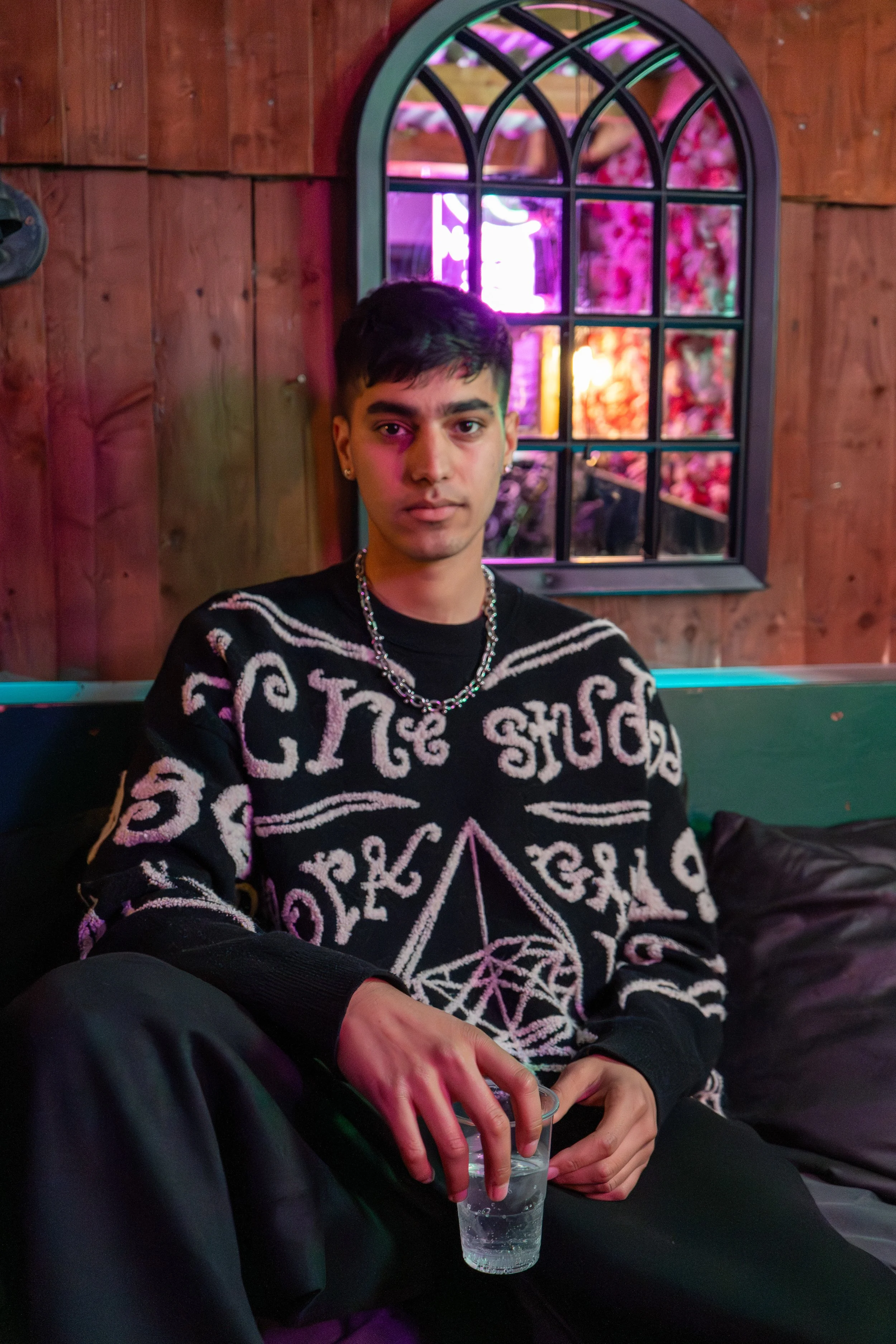 A young man with dark hair, wearing a black sweater with white graphic prints and jewelry, sitting on a black couch in a dimly lit room with wooden walls and a window showing pink and purple neon lights.