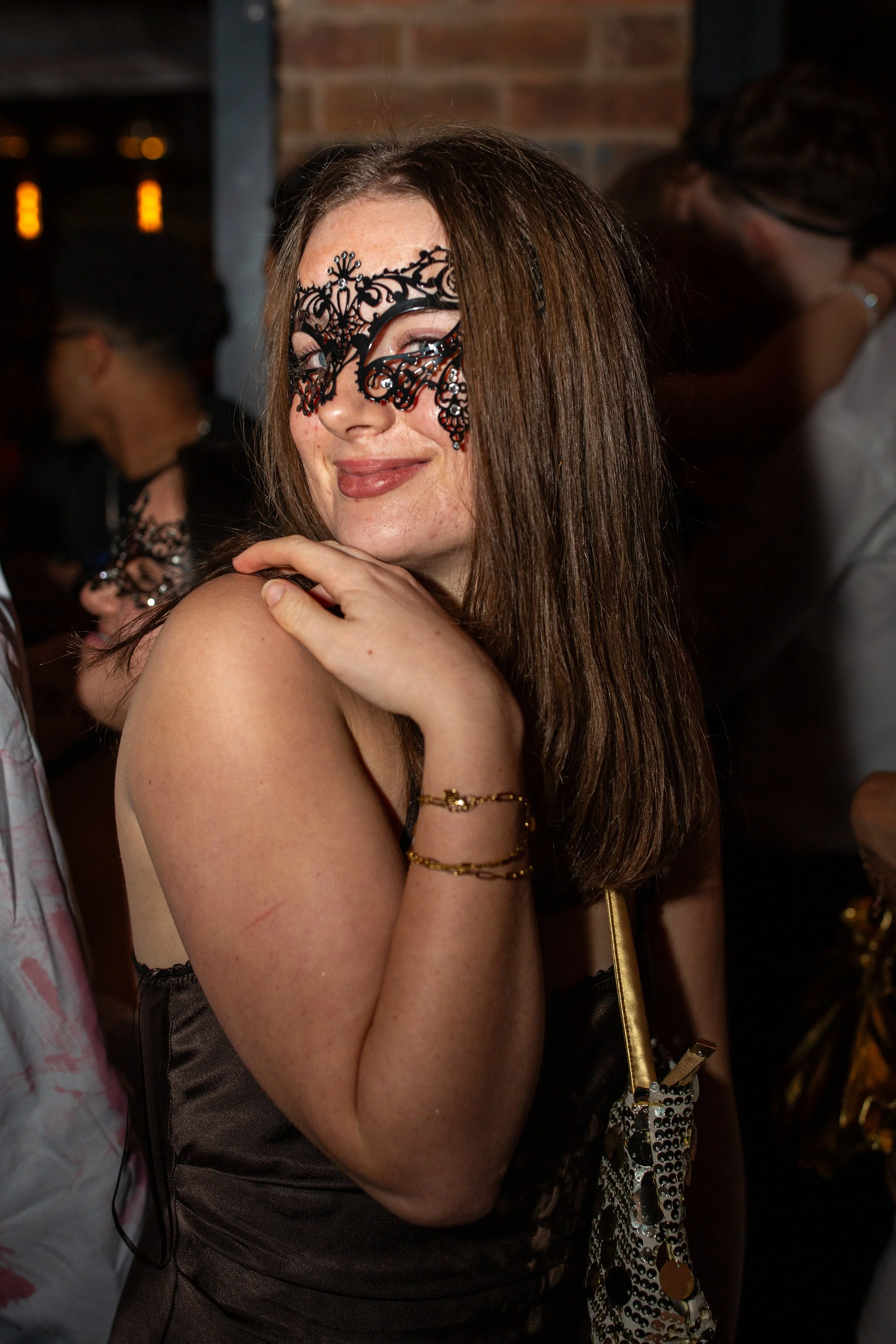 A woman with long brown hair wearing an ornate black lace mask at a social event, smiling and looking at the camera.