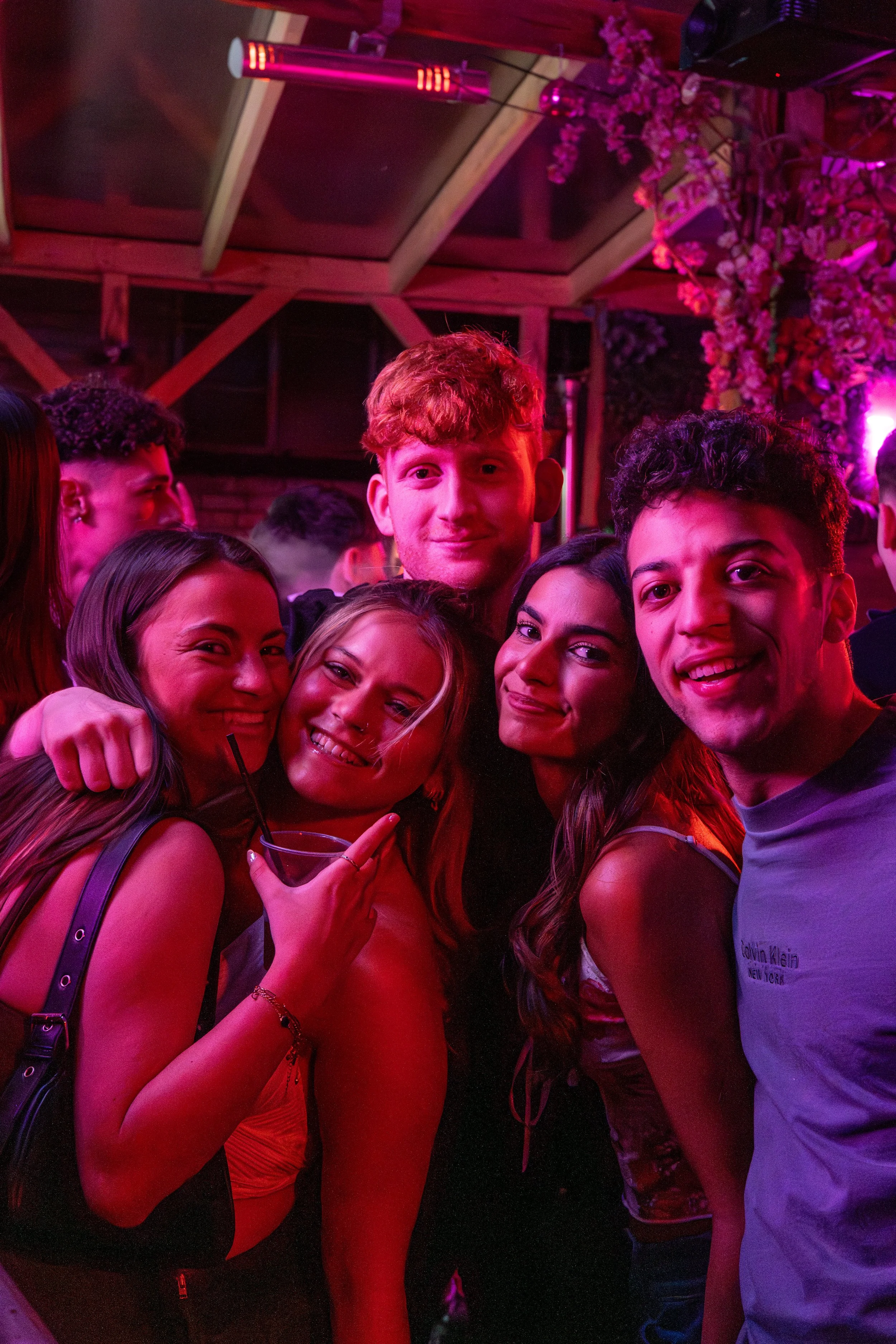 Group of friends posing together at a party or nightclub with pink and purple lighting, some holding drinks, and festive decorations in the background.
