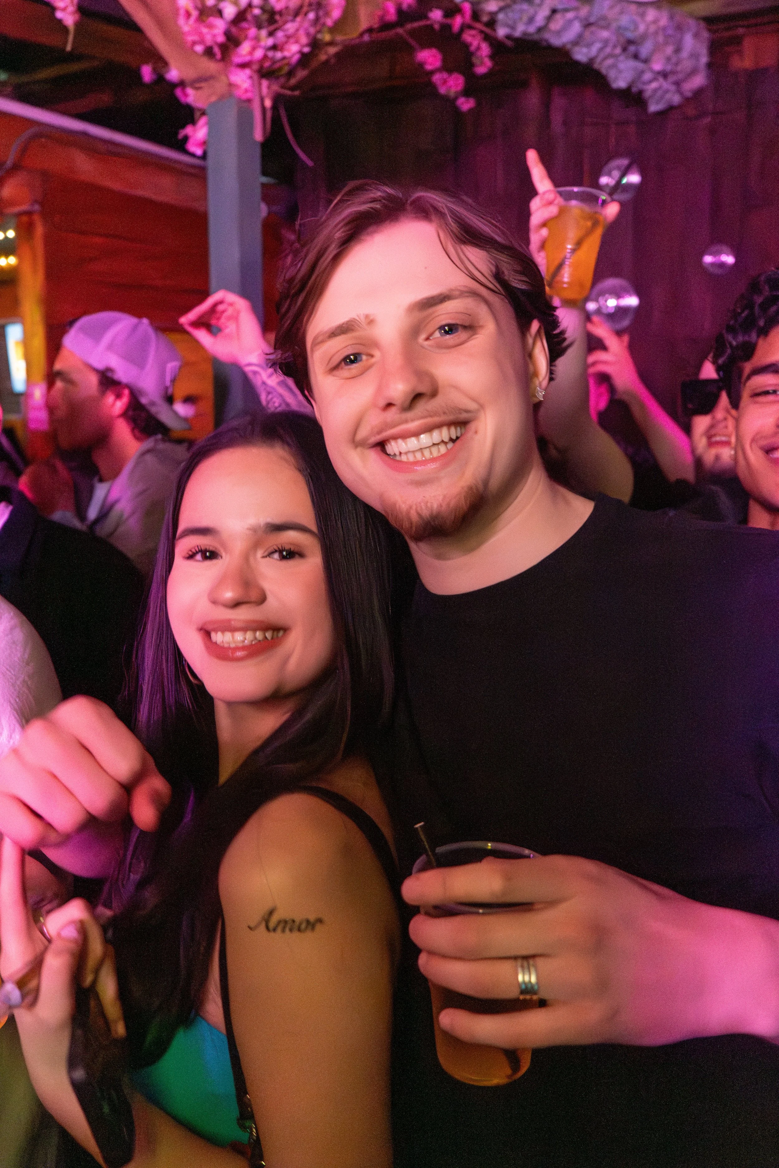 Two young adults smiling at a party or club, surrounded by other people, some holding drinks and dancing.