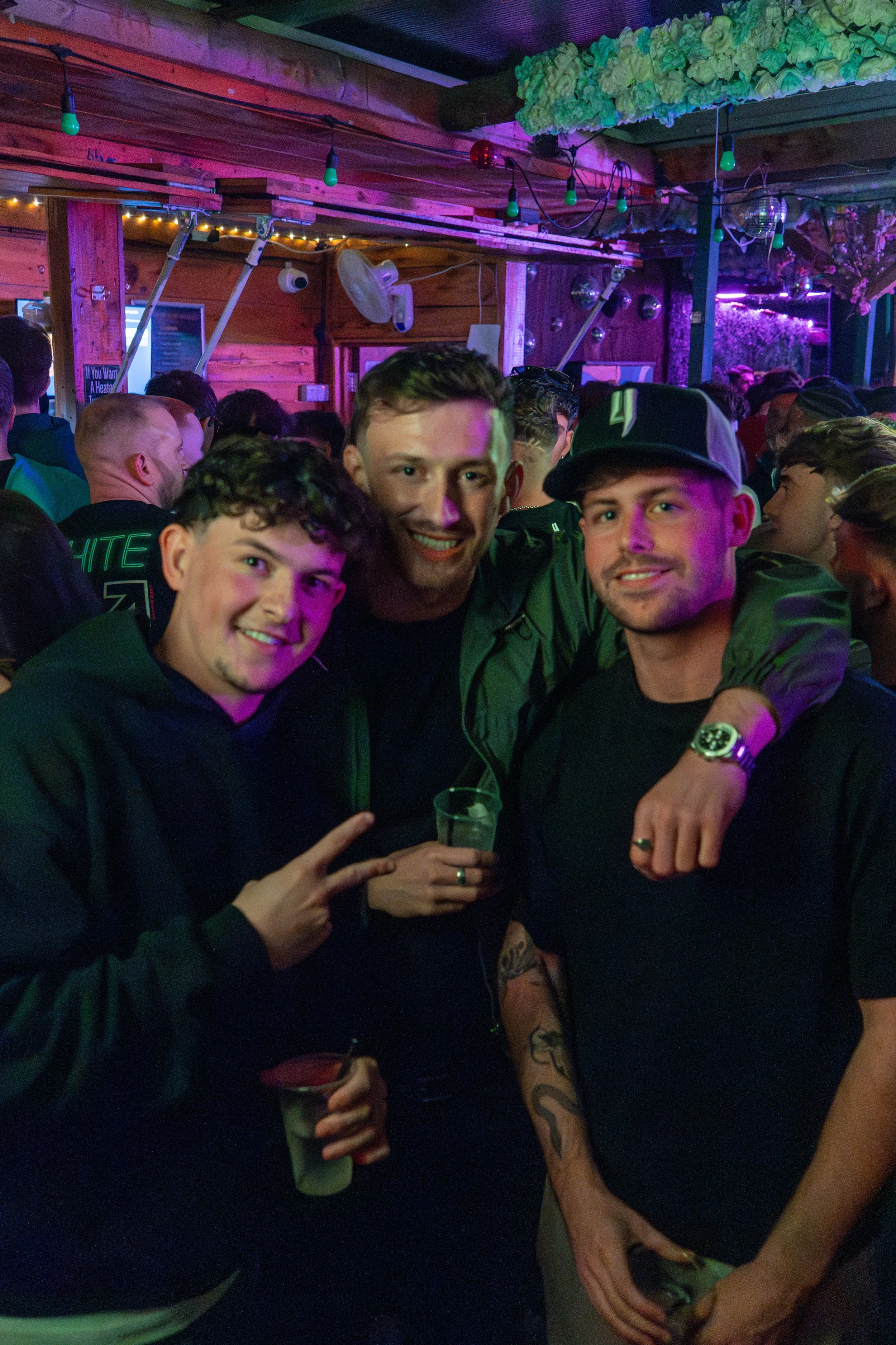Three young men smiling at a party or club with colorful lighting, crowd in the background, and one holding a drink.