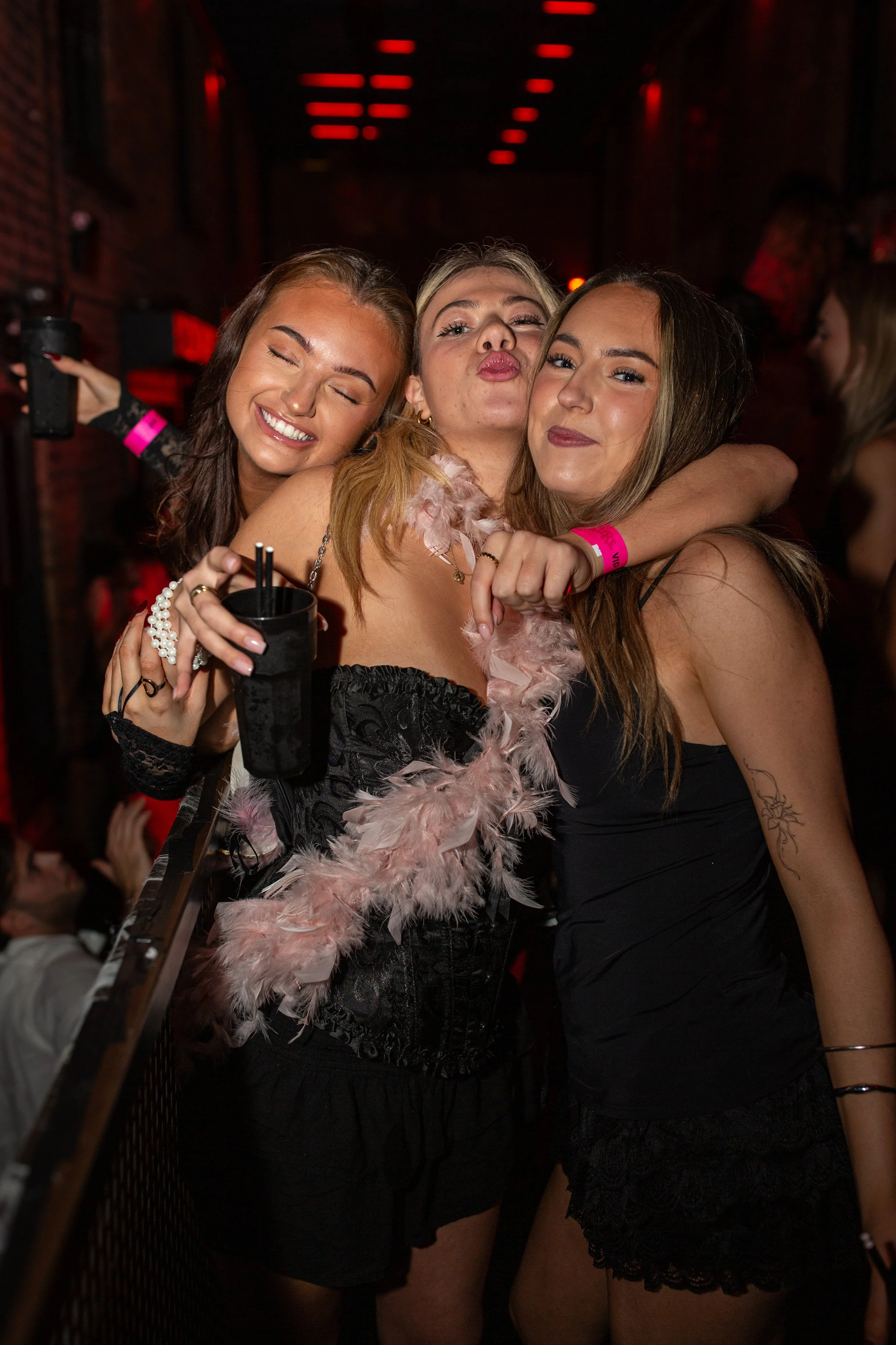 A group of three young women hugging and smiling at a party or club with red lighting in the background.