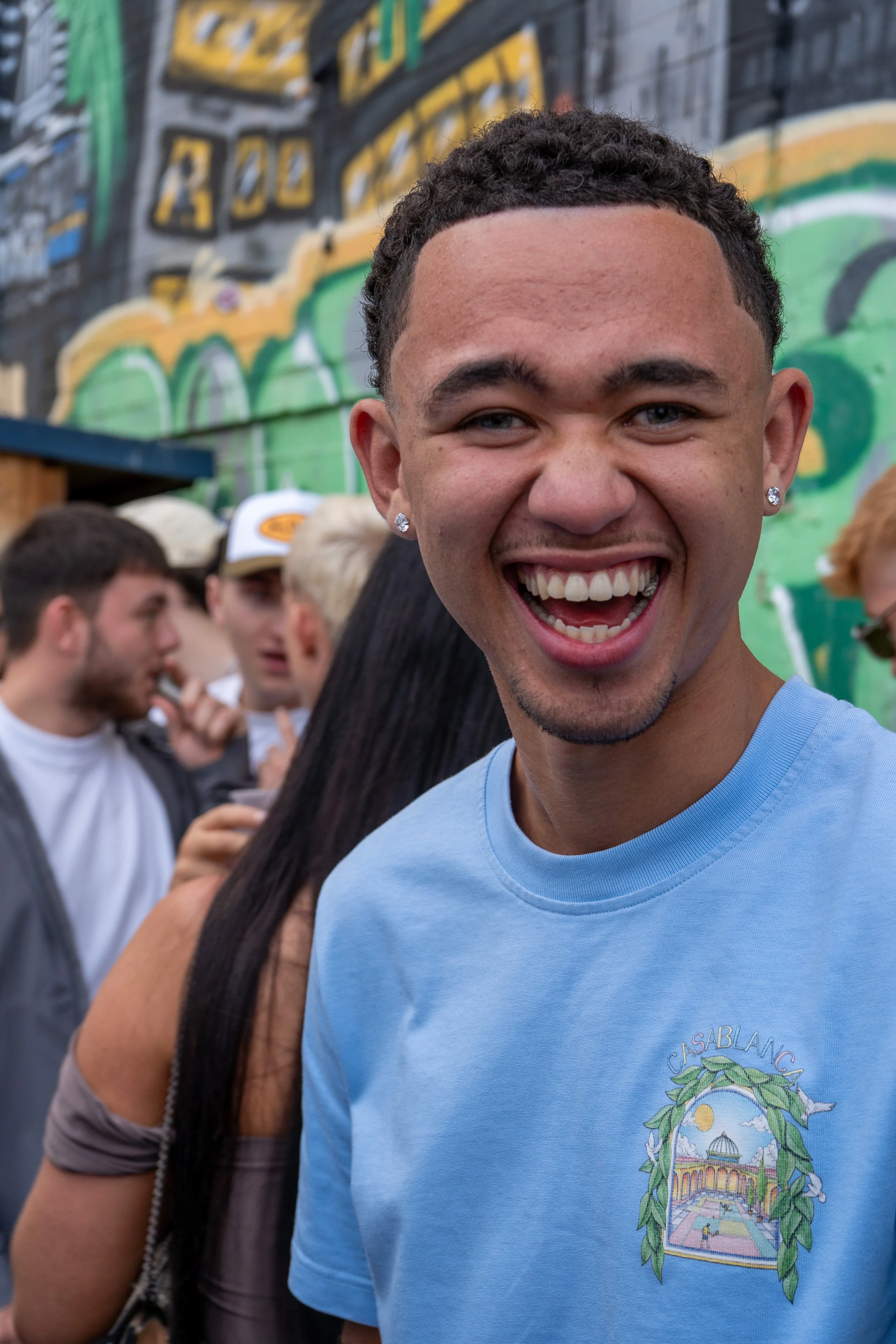 A young man with short curly hair, earrings, and a bright smile wearing a light blue T-shirt with a colorful graphic design stands in front of a crowd at an outdoor event with a graffiti-covered wall in the background.