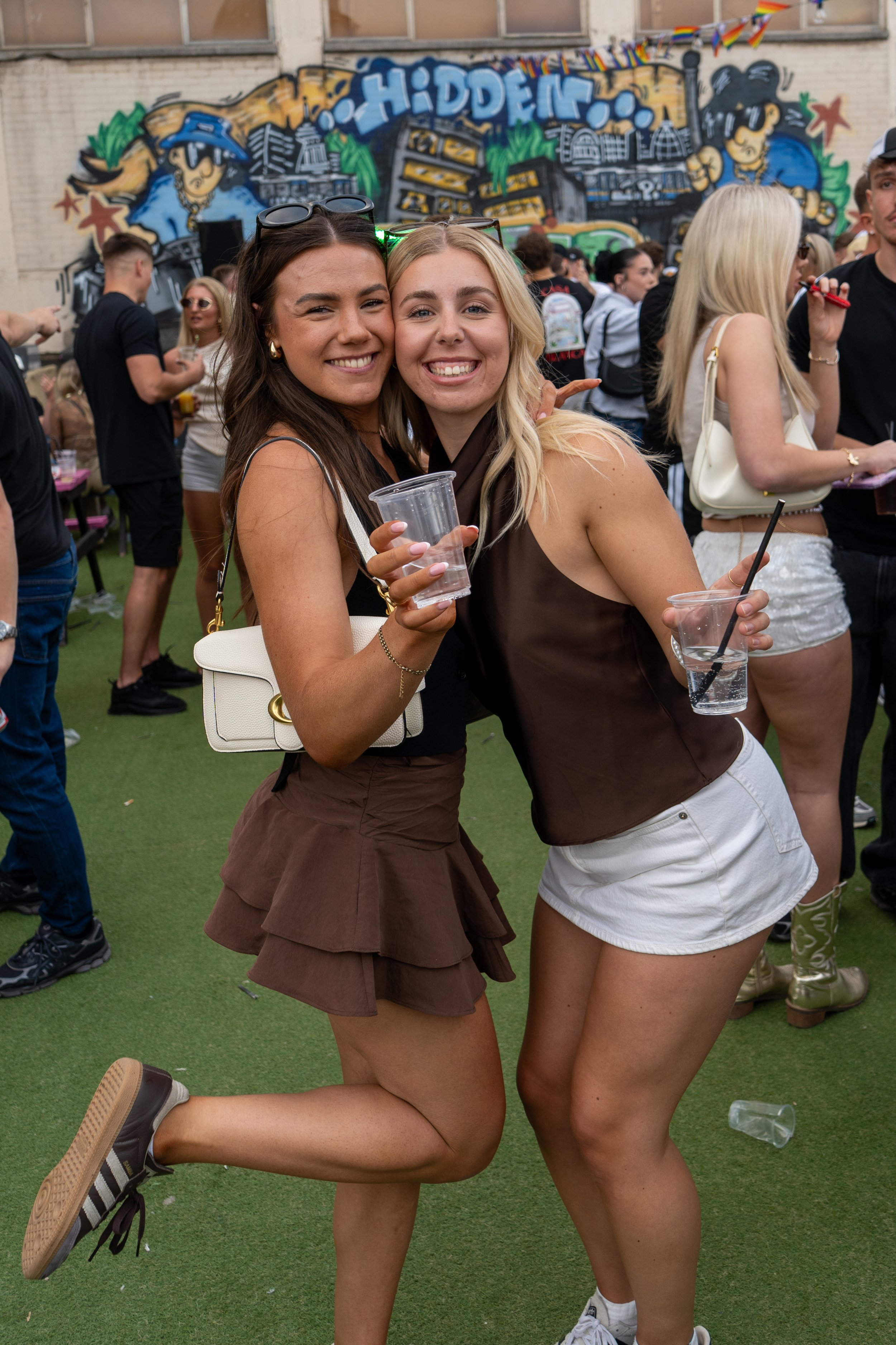 Two young women smiling, hugging, and holding drinks at an outdoor event with a graffiti wall and other people in the background.