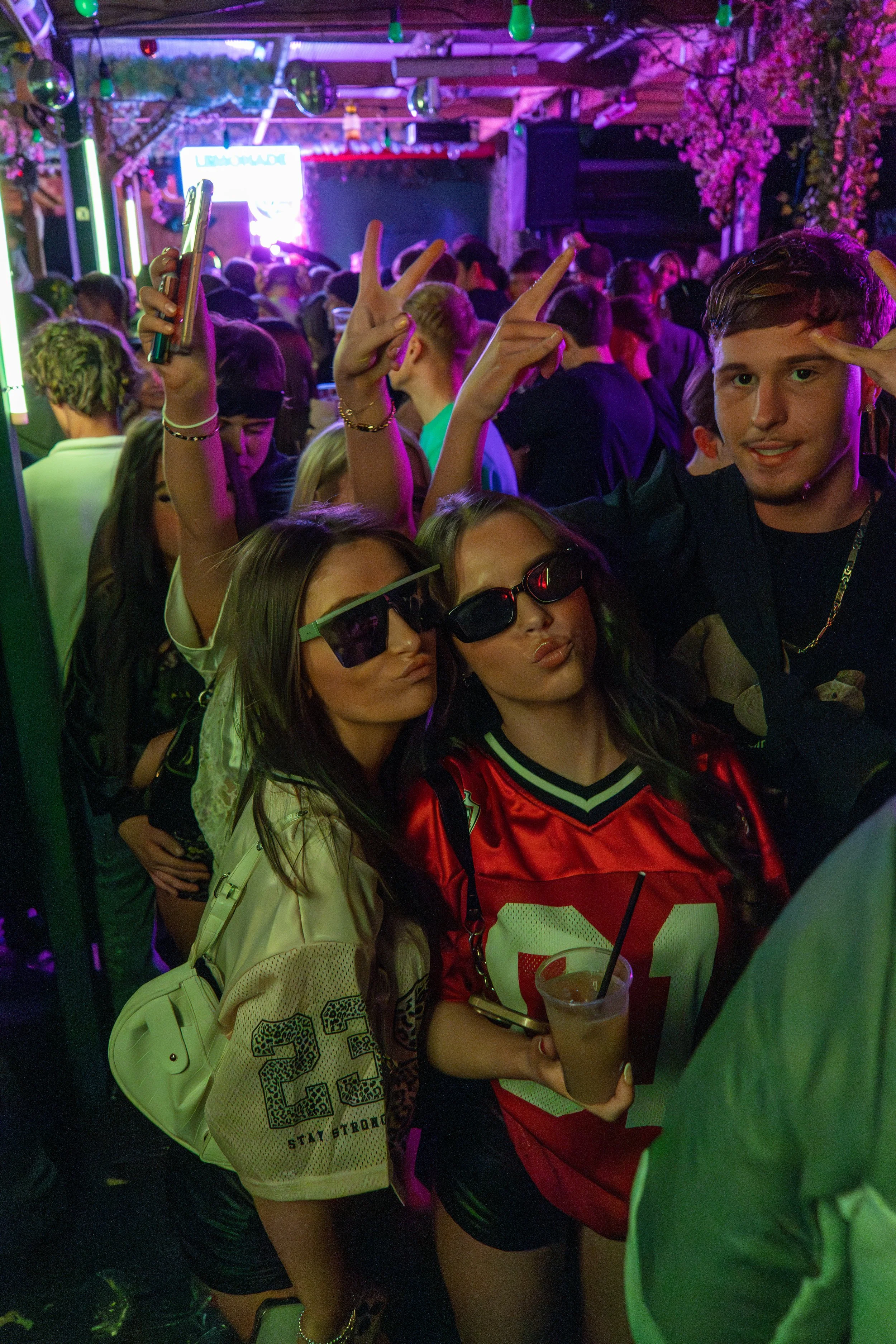 Group of young people at a crowded indoor party with neon and colorful lights, some wearing sunglasses, taking selfies, and mingling.