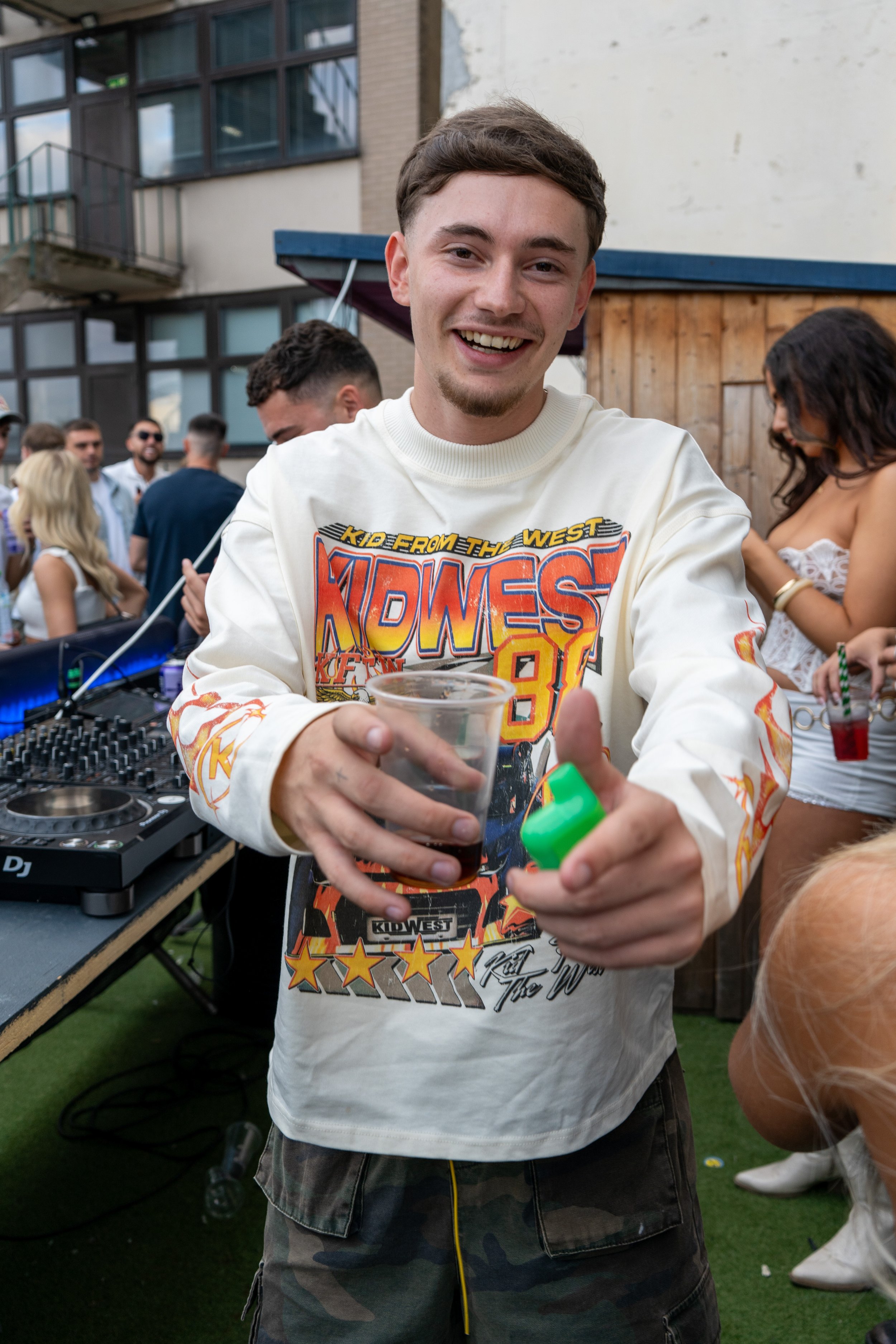 A young man with short brown hair and light facial hair smiling at the camera while holding a drink and a green object at a lively outdoor party or gathering.