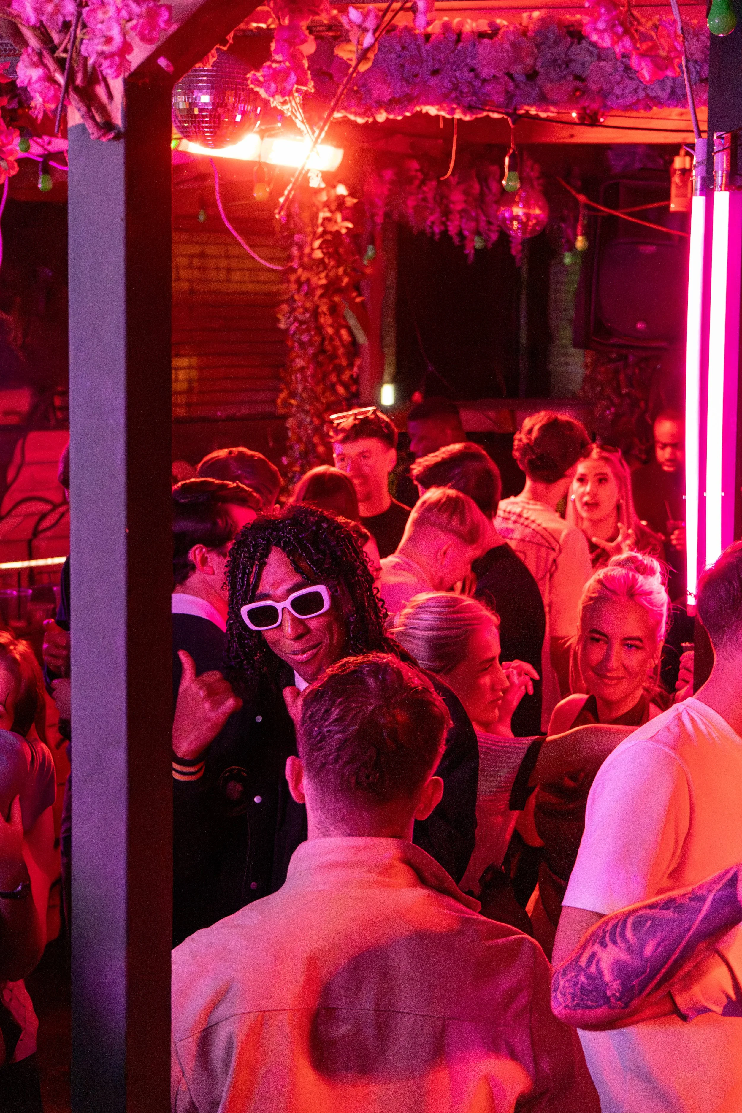 People socializing and dancing at a vibrant nightclub, illuminated by red and pink lights, with floral decorations and neon signs.