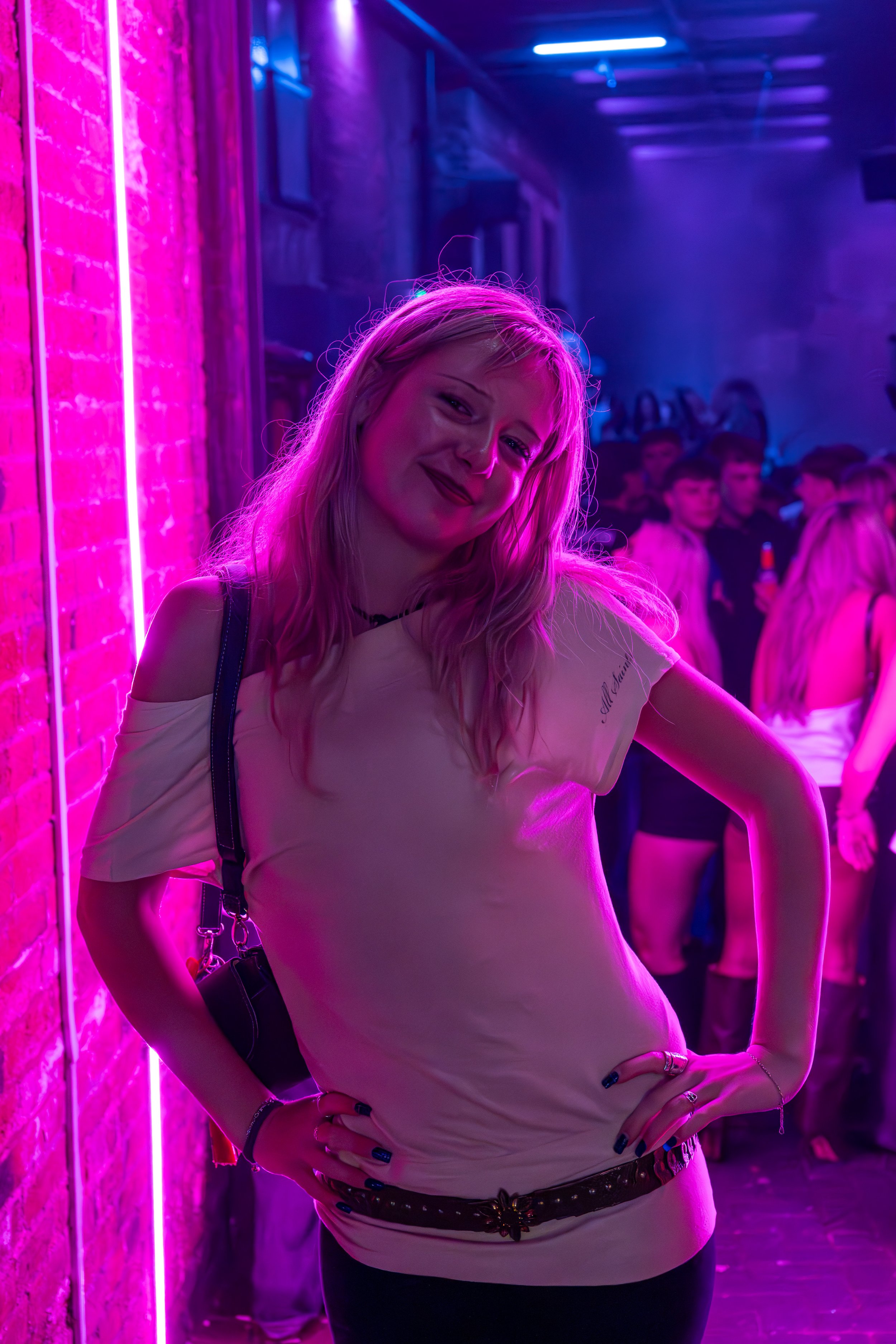 A young woman with blonde hair smiling at a party or club with purple neon lighting, standing against a brick wall with other people in the background.