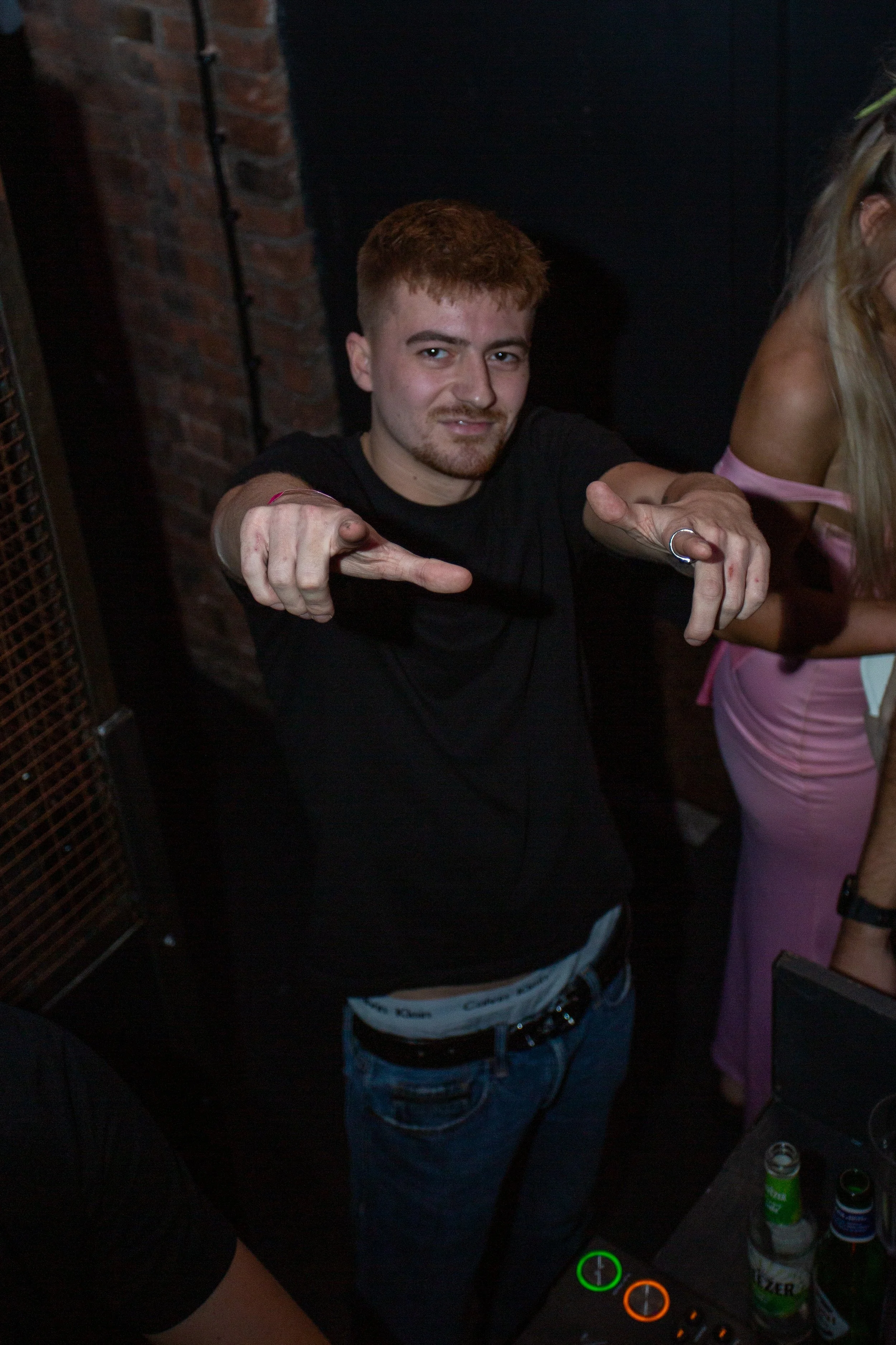 A young man with short red hair and a beard pointing towards the camera at a party or social gathering, standing in front of a brick wall with drinks on a table nearby.