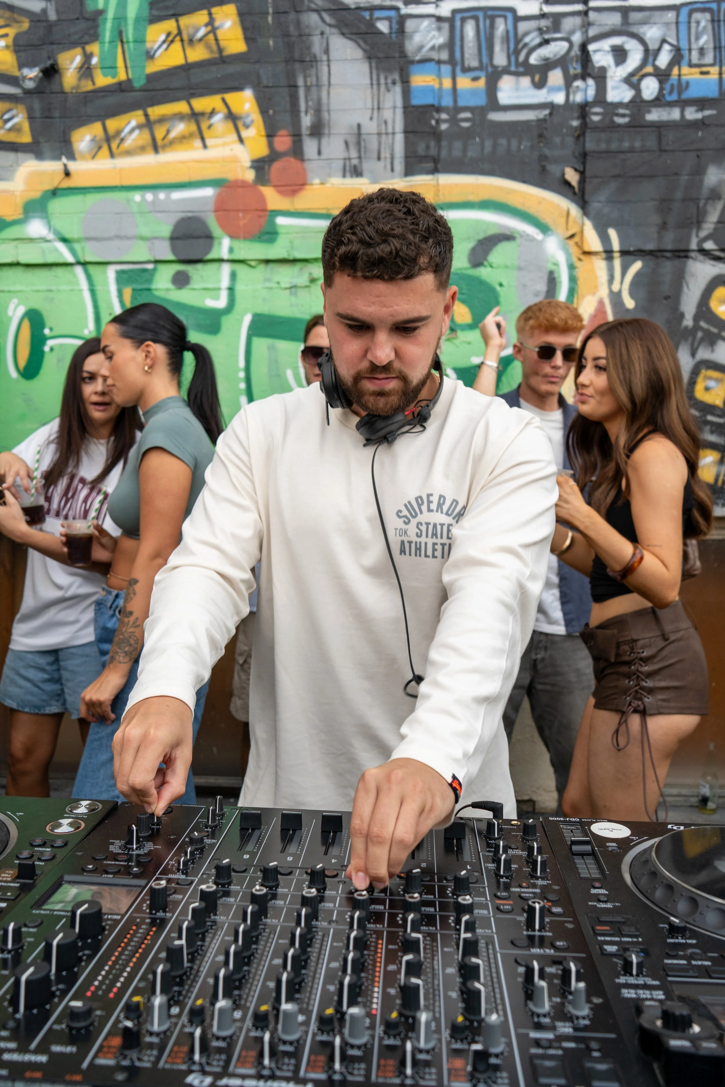 A male DJ wearing a white long-sleeve shirt and headphones around his neck mixing music at his DJ equipment. Behind him, a group of people are socializing in front of a graffiti-covered brick wall.