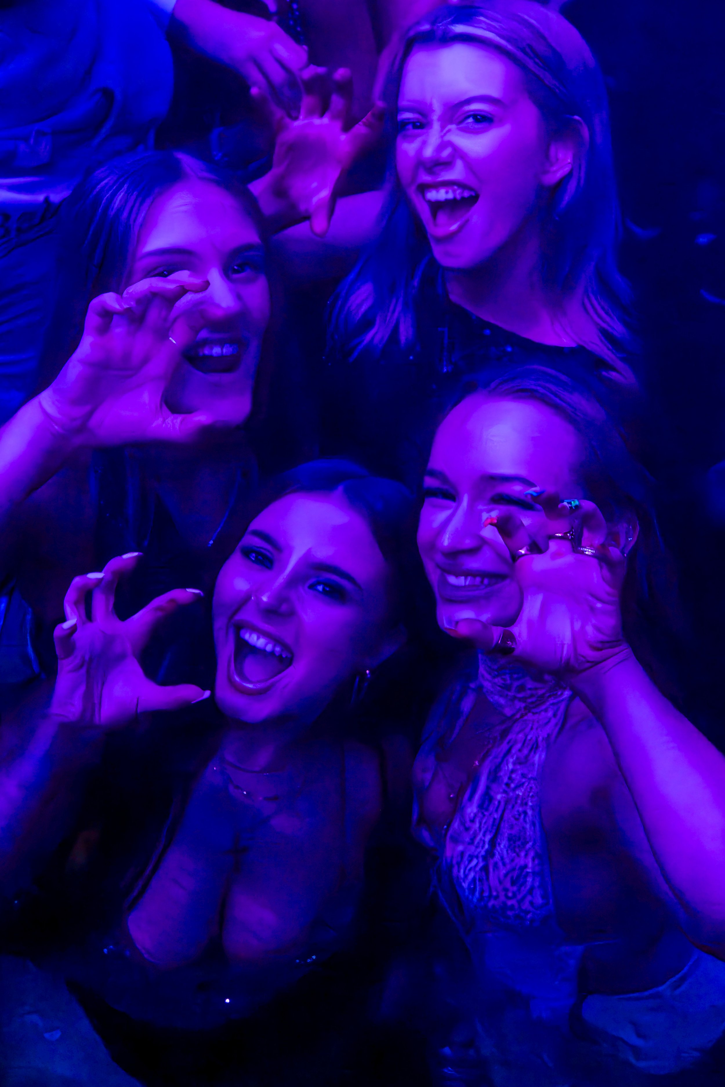 Four young women smiling and making hand gestures in a dimly lit, purple-hued setting, likely a nightclub or party.