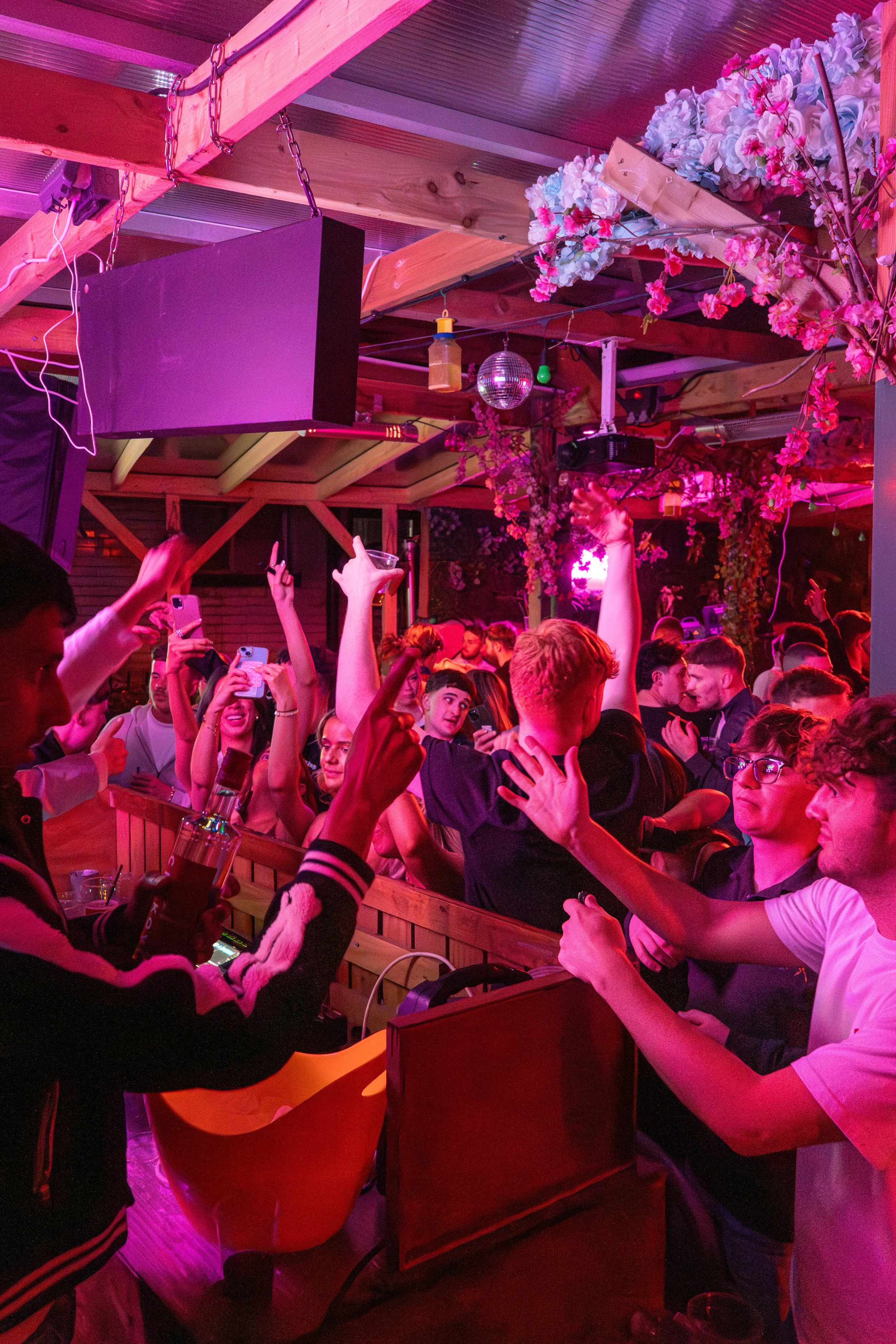 Crowd of young people partying, dancing, and taking photos at a lively nightclub with pink and purple lighting, floral decorations, and wooden interior elements.