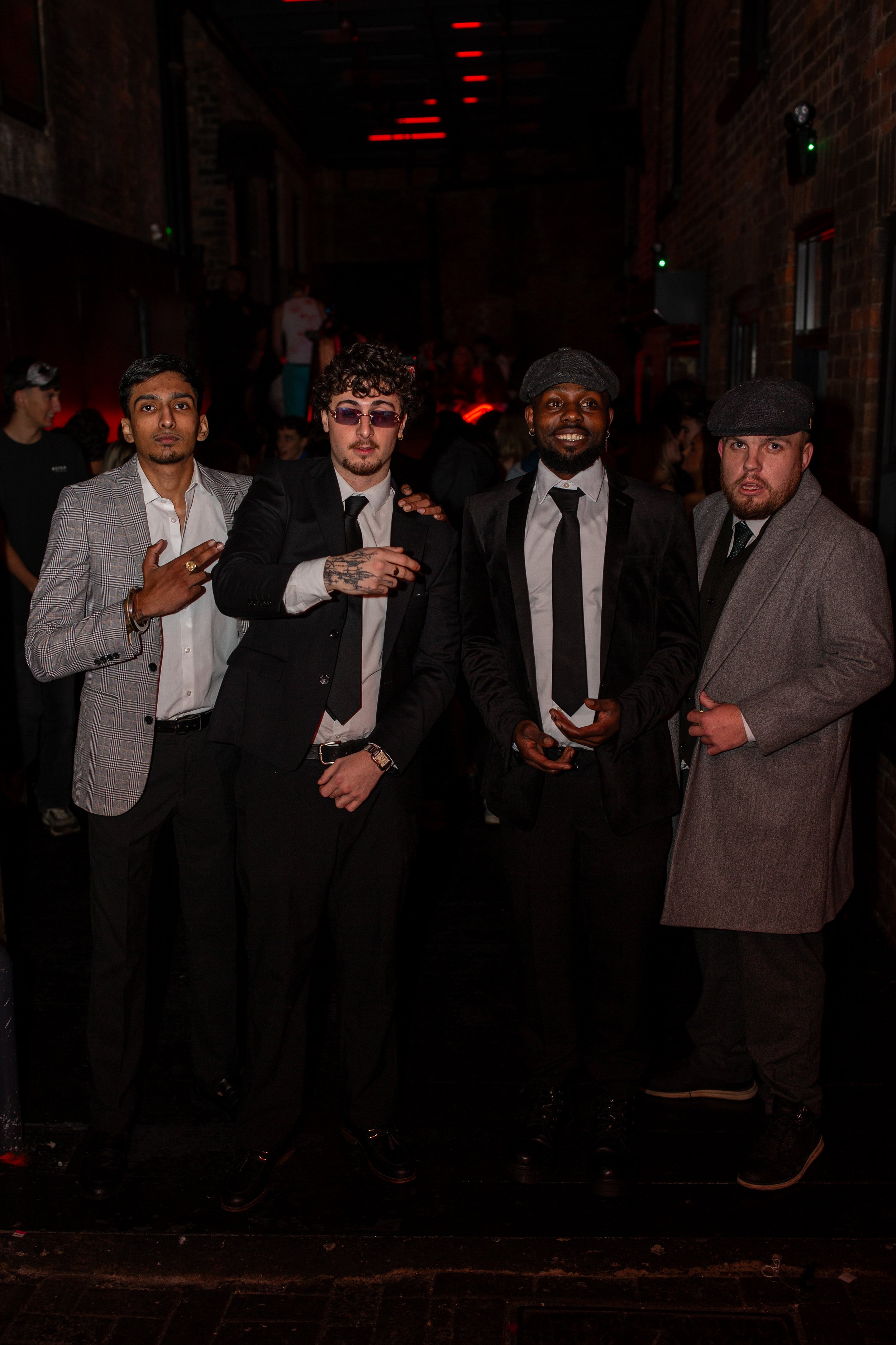 Four men standing together at a nighttime party, dressed in suits, with a dimly lit venue behind them.
