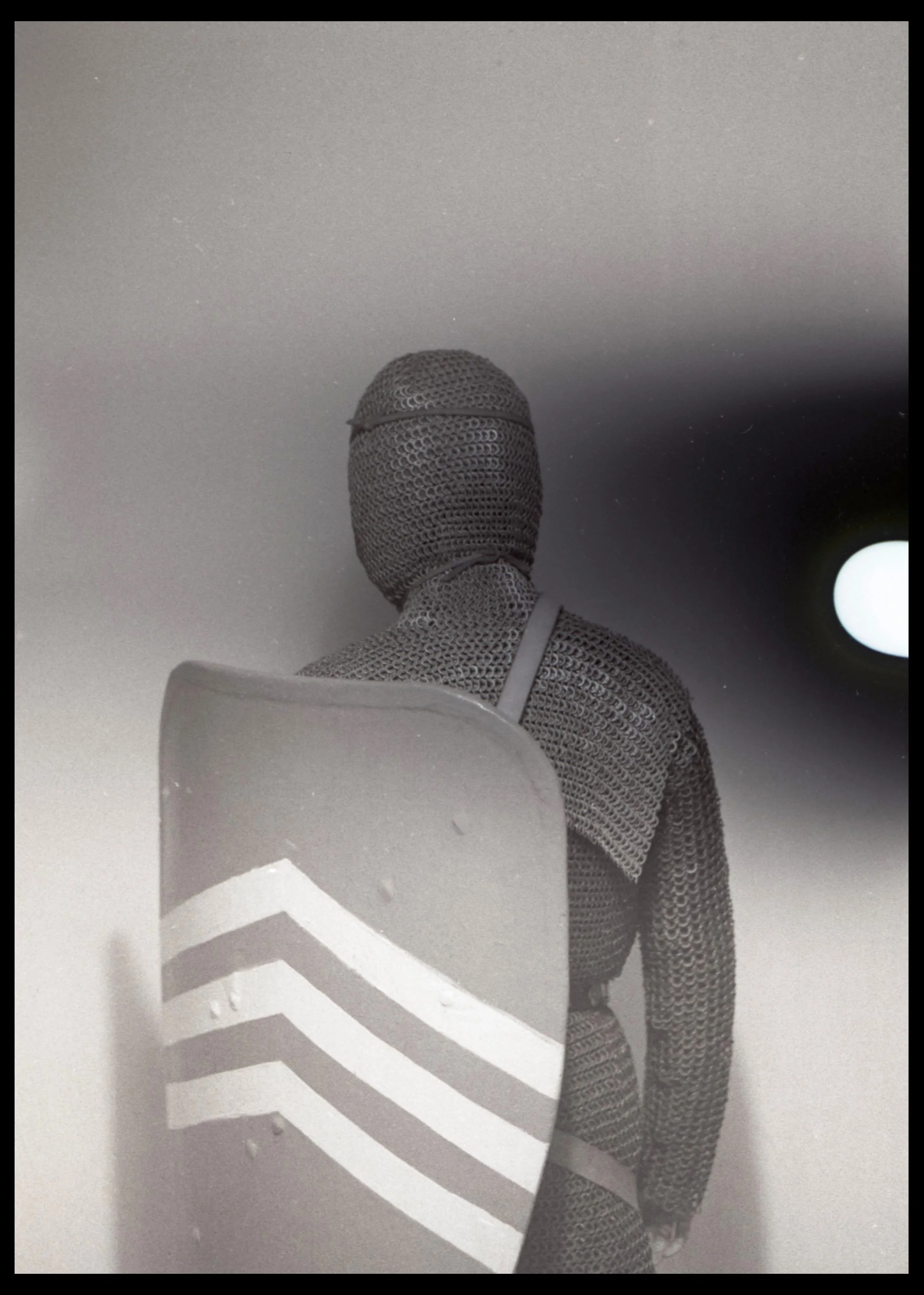 A mannequin dressed in chainmail armor holding a shield with three white stripes, standing against a plain wall with a round light fixture in the background.