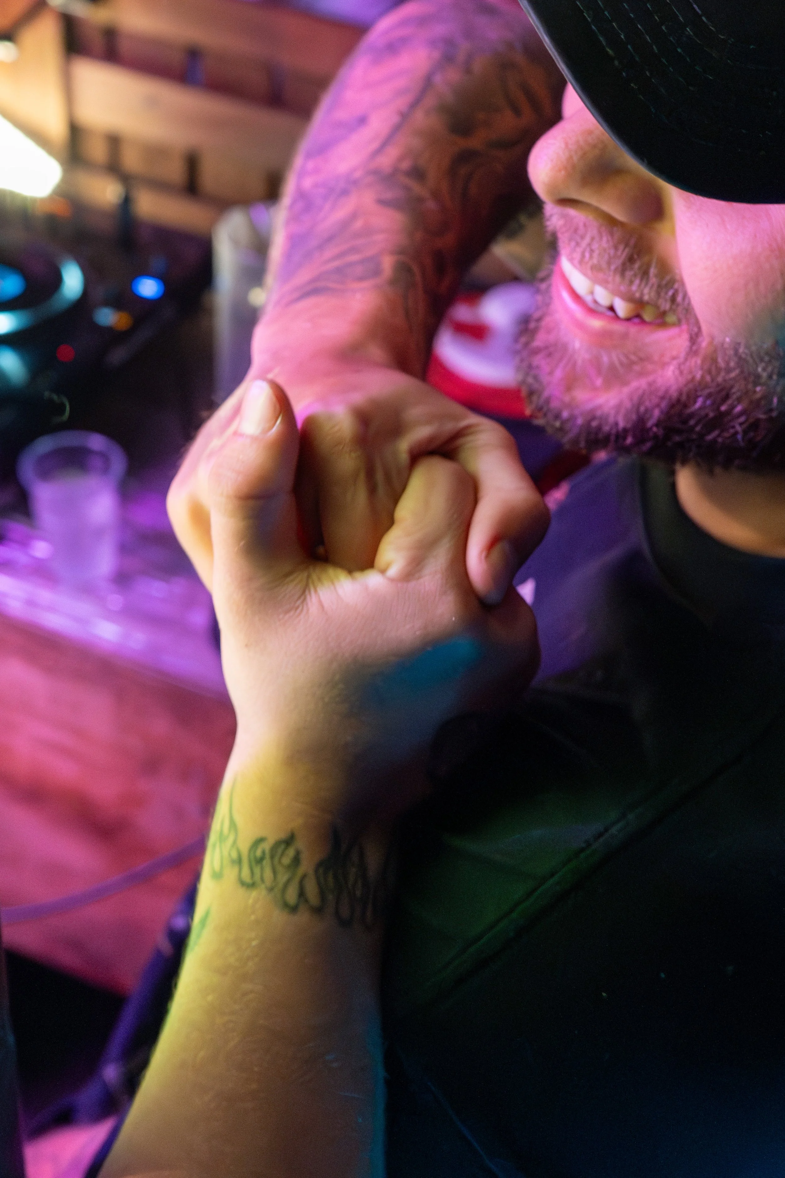 A person with tattoos and a beard smiling and engaging in a fist bump with another person, near a DJ setup with turntables in the background.
