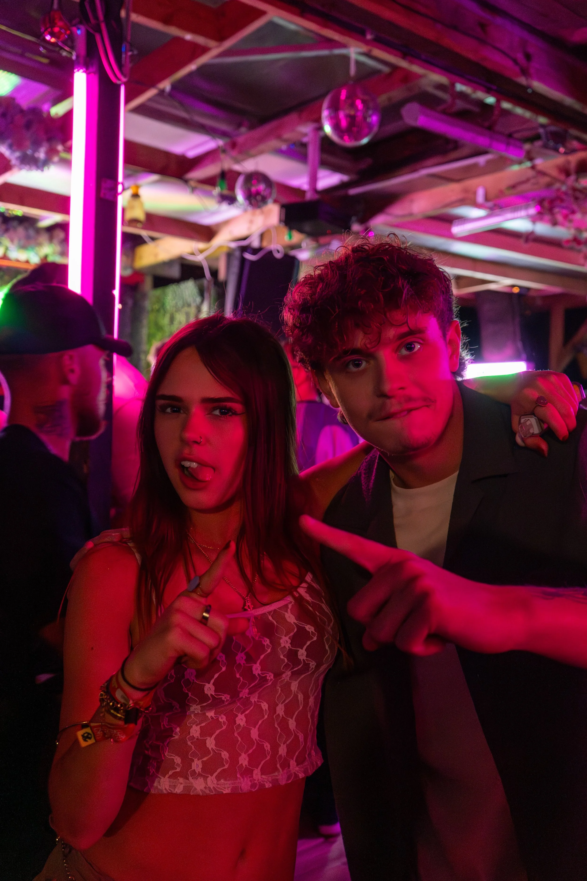 Young woman and man posing at a party with pink and purple neon lighting, disco balls, and people in the background.