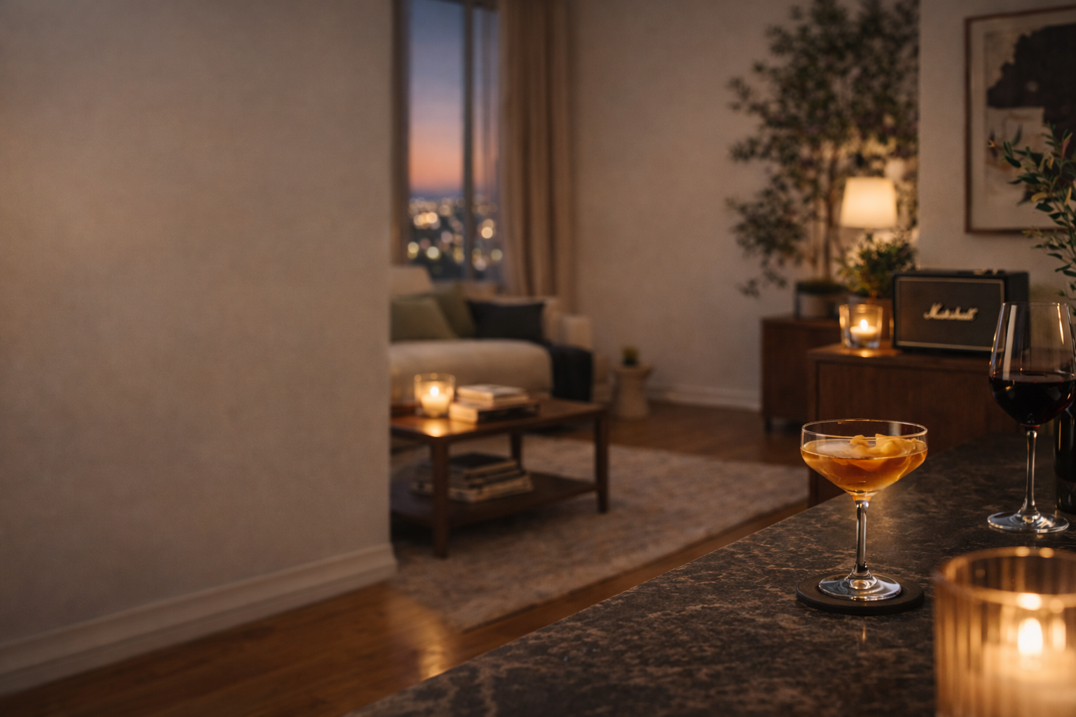 A cozy living room scene viewed from a kitchen counter. The counter has a glass of amber-colored drink and a glass of red wine. In the background, there is a sofa with cushions, a coffee table with candles and books, a window showing a cityscape at sunset, and ambient lighting from lamps and candles creating a warm atmosphere.