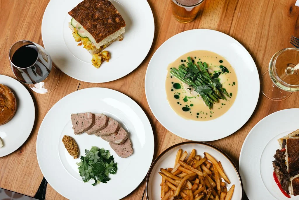An overhead view of a wooden table with various plates of food, including sliced pate with mustard and greens, a plate of fries, a slice of brown bread, green asparagus in creamy sauce, and glasses of wine and beer.
