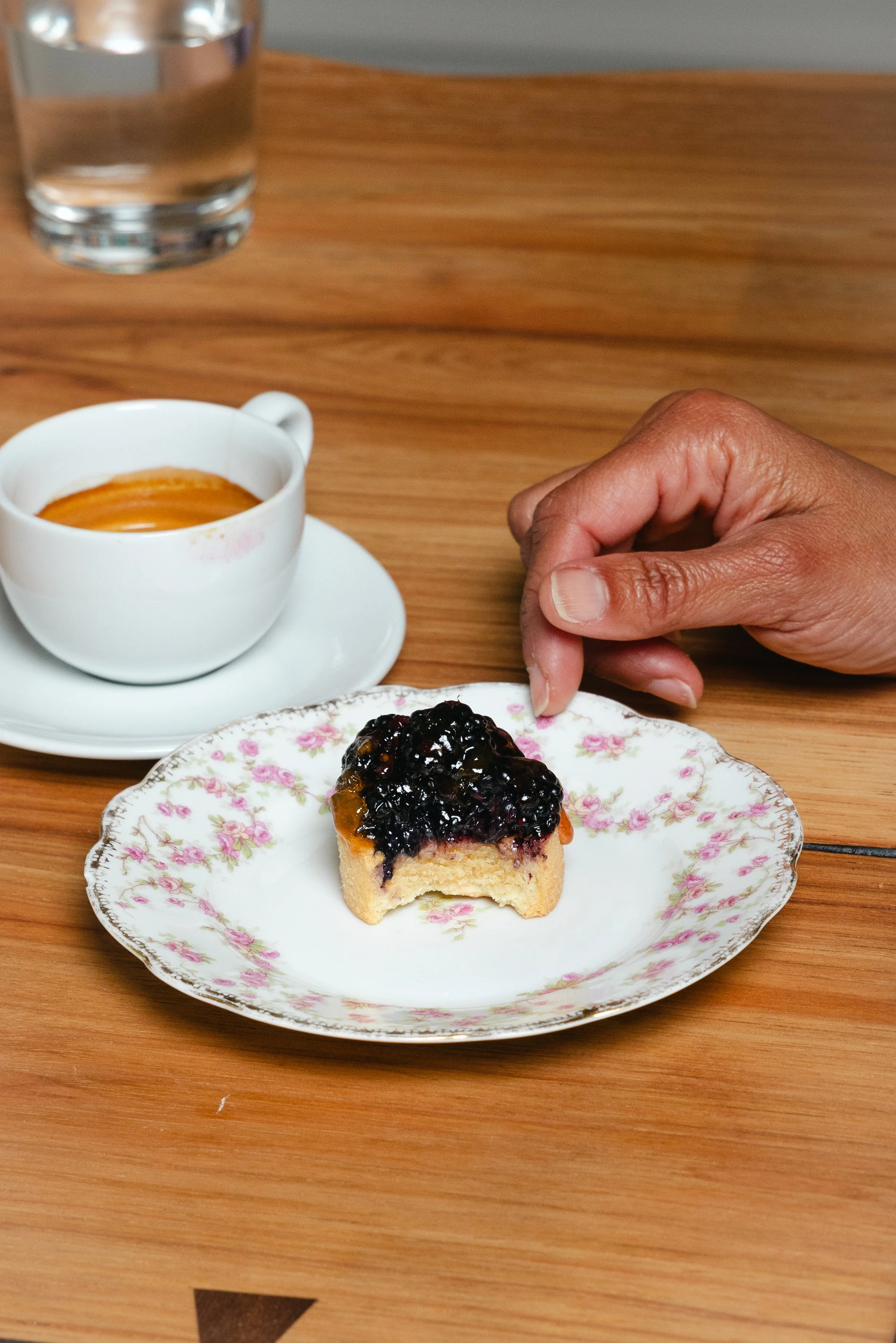 A dessert topped with blackberries on a floral-patterned plate, a shot of espresso, and a glass of water on a wooden table.