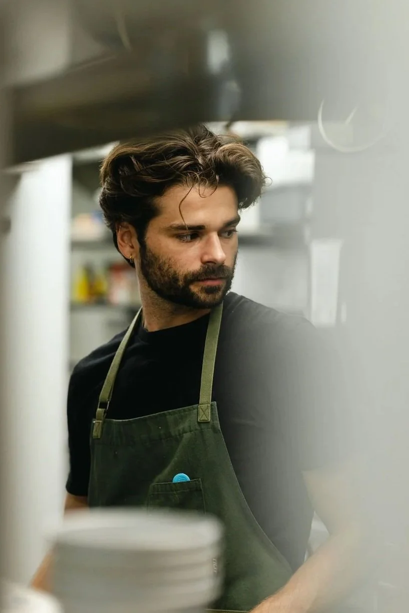 A man wearing a black shirt and a green apron, standing in a kitchen with shelves and bottles in the background, looking to the side with a focused expression.