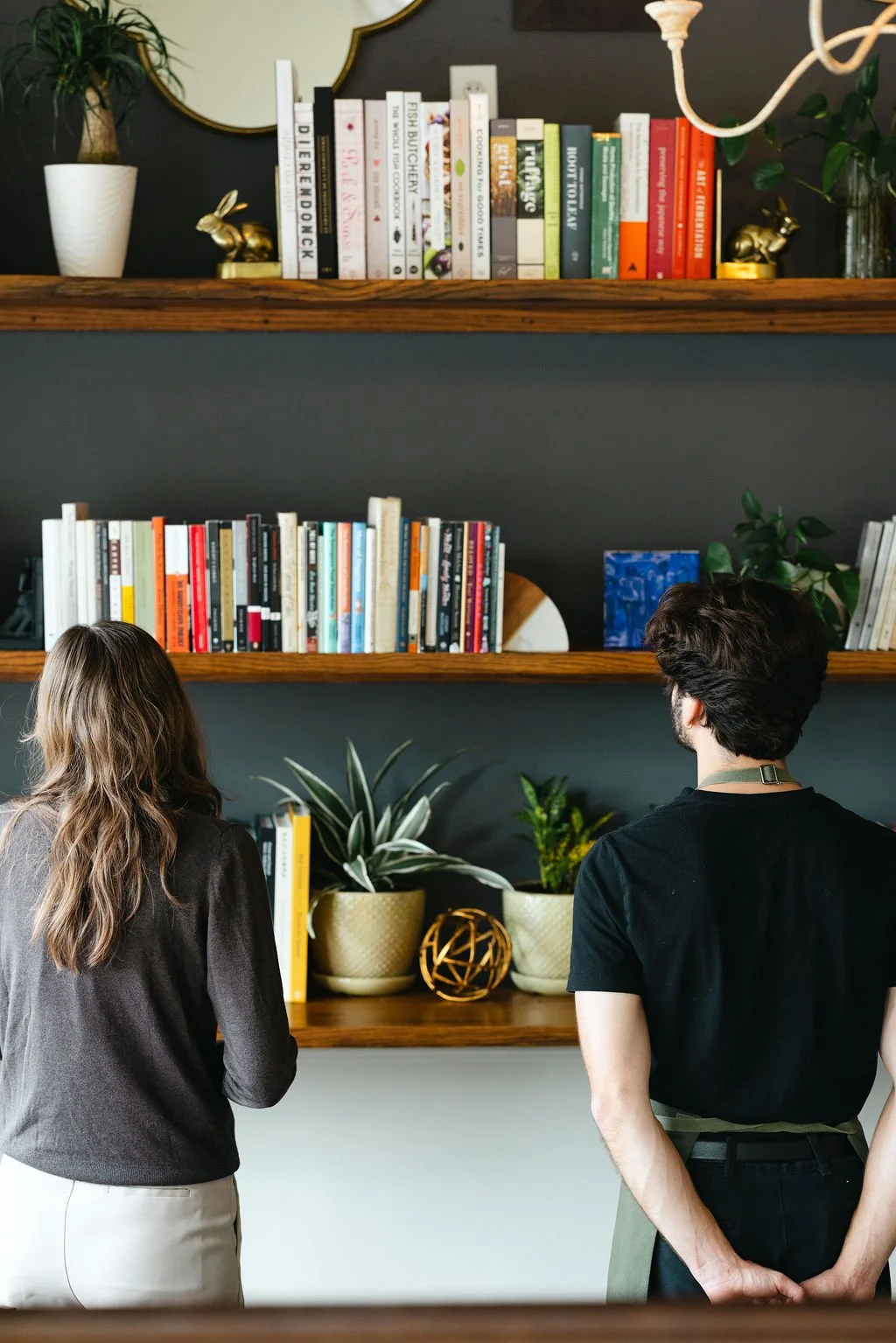 Two people, a woman with long wavy hair wearing a brown sweater and a man with dark hair wearing a black t-shirt, stand in front of wooden shelves filled with books and decorative plants.