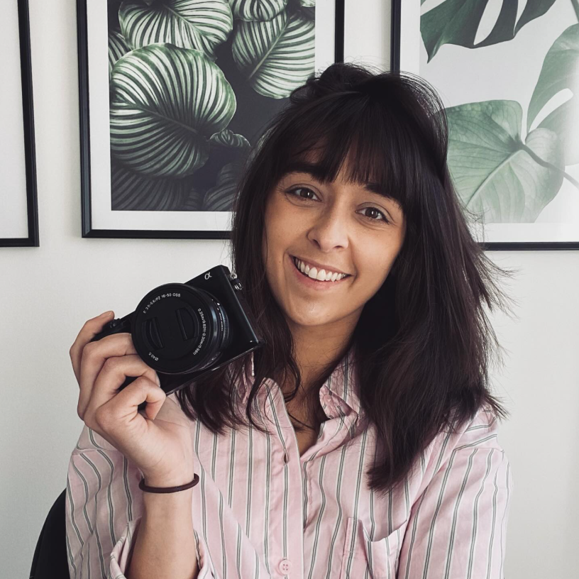 A woman with shoulder-length dark hair and bangs smiling while holding a camera, sitting in front of framed botanical artwork featuring green leaves.