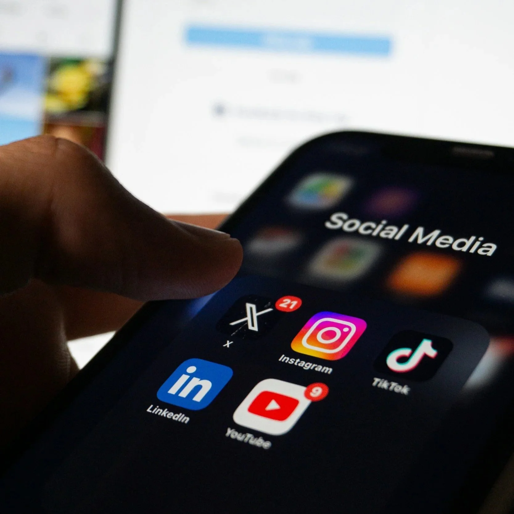 Close-up of a smartphone screen displaying social media app icons including Instagram, TikTok, LinkedIn, Facebook, YouTube, and Twitter, with a person's thumb visible interacting with the screen.