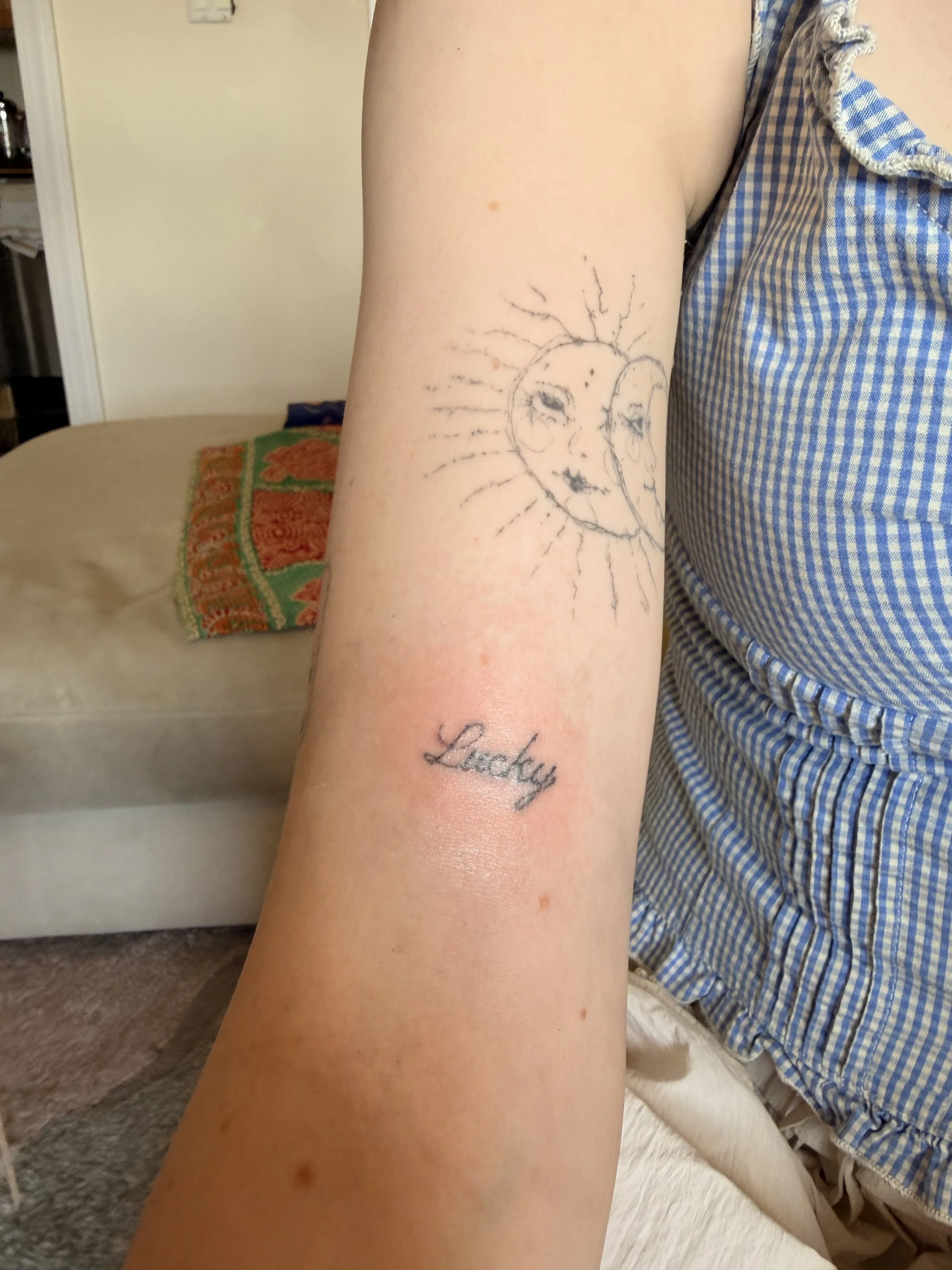 A person's inner forearm with a tattoo of a smiling sun with a face and a second face looking down, with  the word "Lucky" tattooed below.