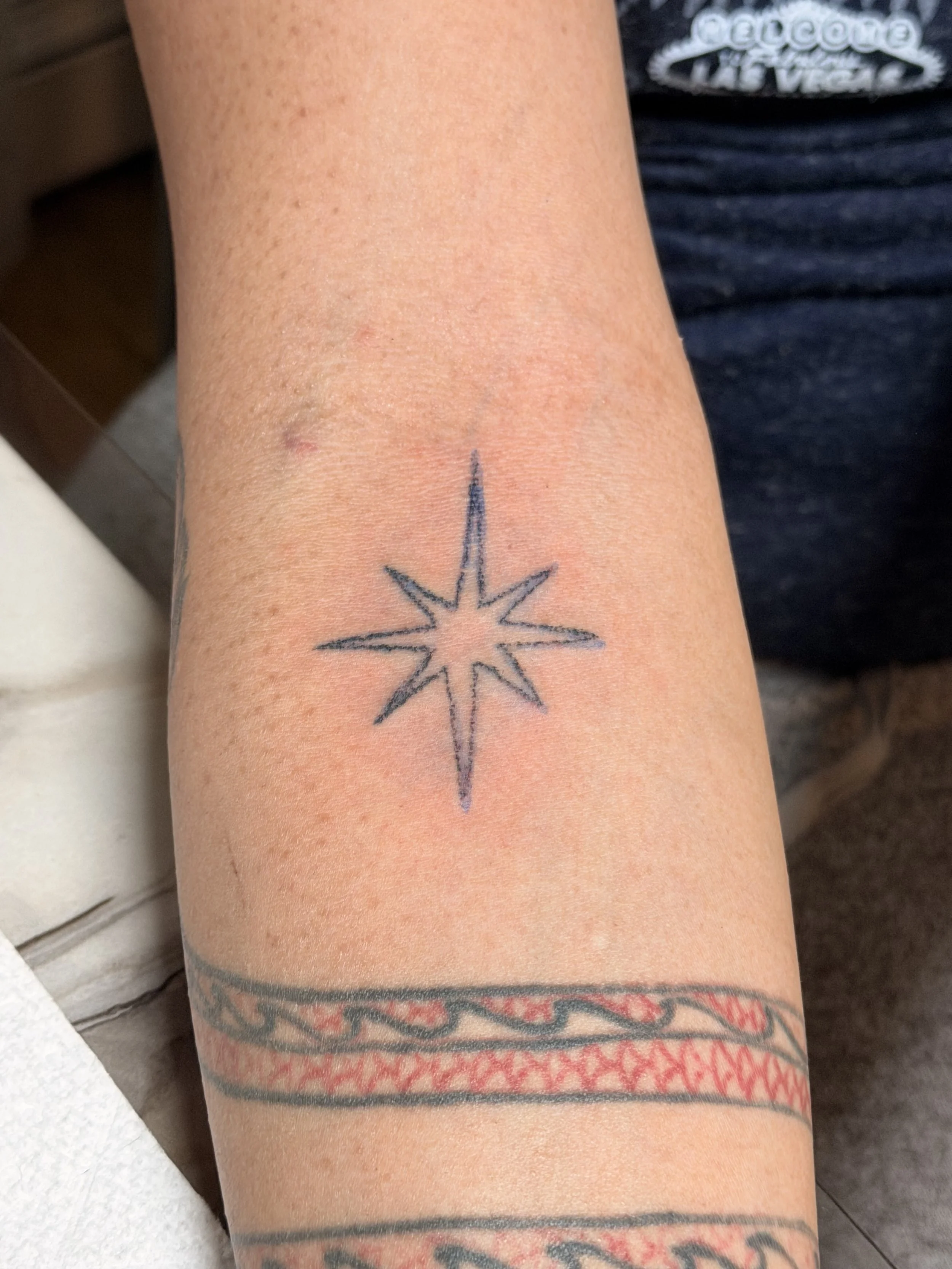 A person's arm with a small tattoo of a star in a compass-like design on the inner forearm, with various additional tattoos visible on other parts of the arm.