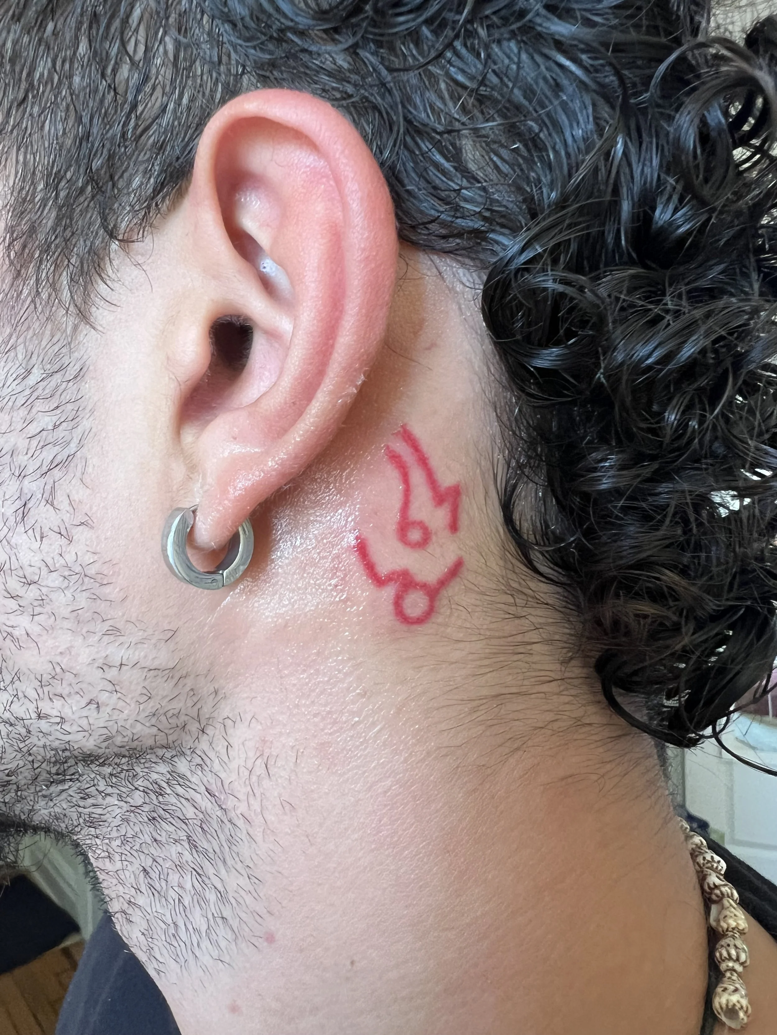 Close-up of a person's left ear with a hoop earring, curly dark hair, and a red tattoo or face paint on the neck.