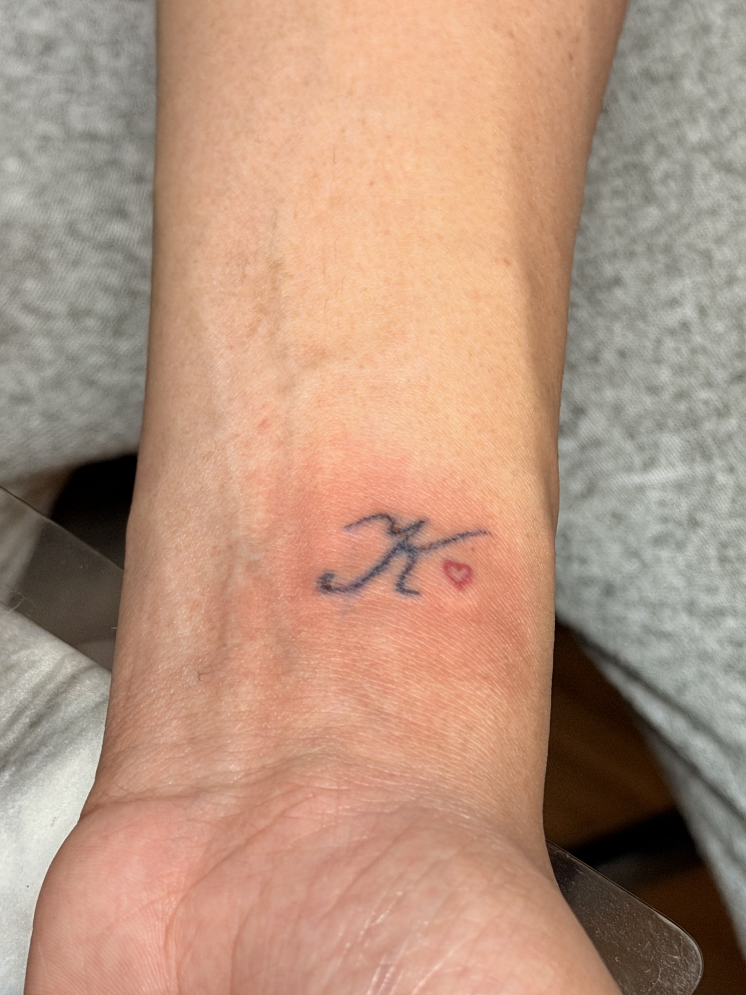 Close-up of a person's wrist with a small tattoo of the letter 'n' in cursive, next to a tiny red heart, and part of a bandage or plaster on the wrist.