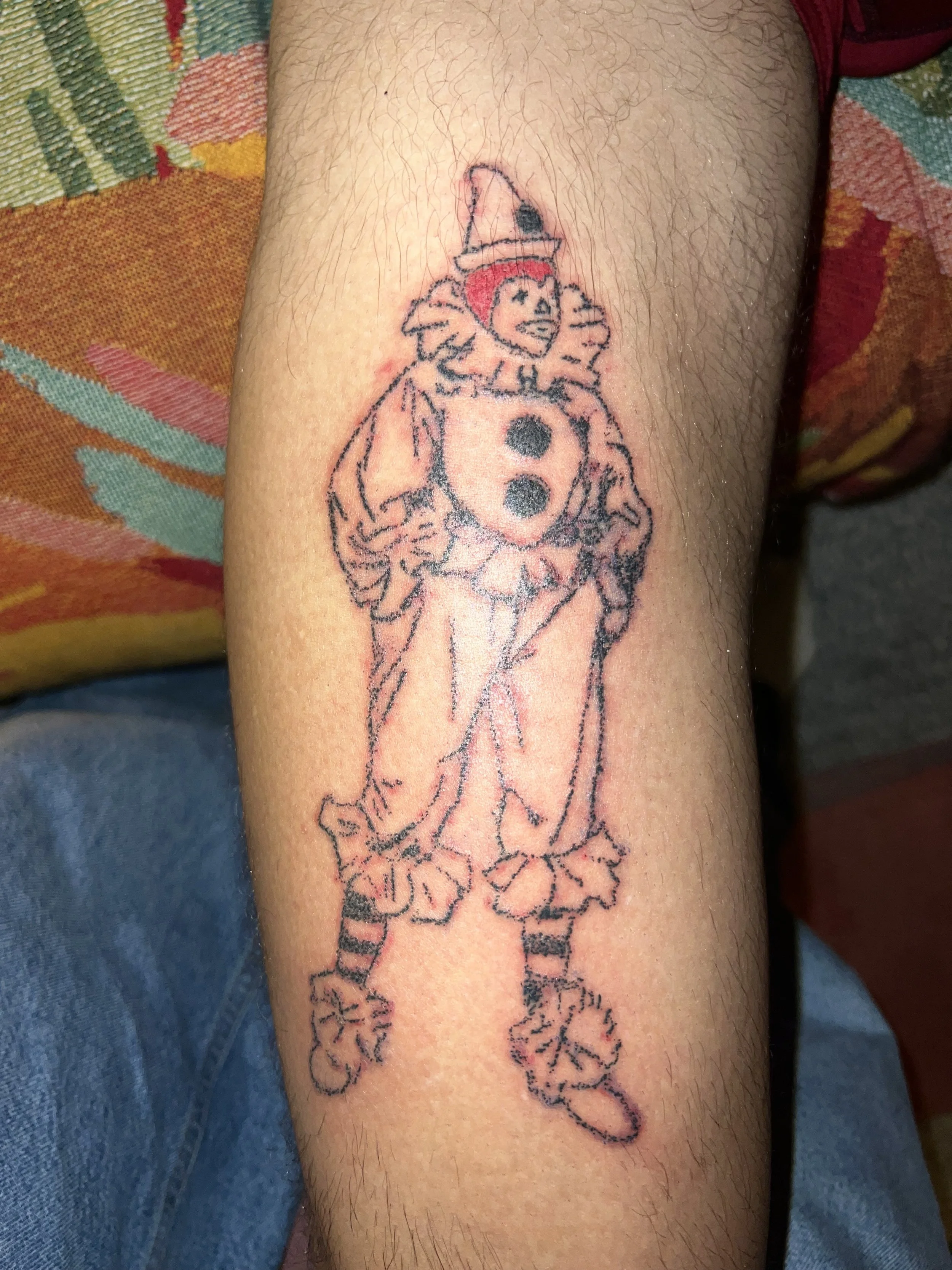 A tattoo of a clown figure on a person's arm, with a pointed hat, a red nose, and a costume with buttons and ruffles, holding a clown's hand.