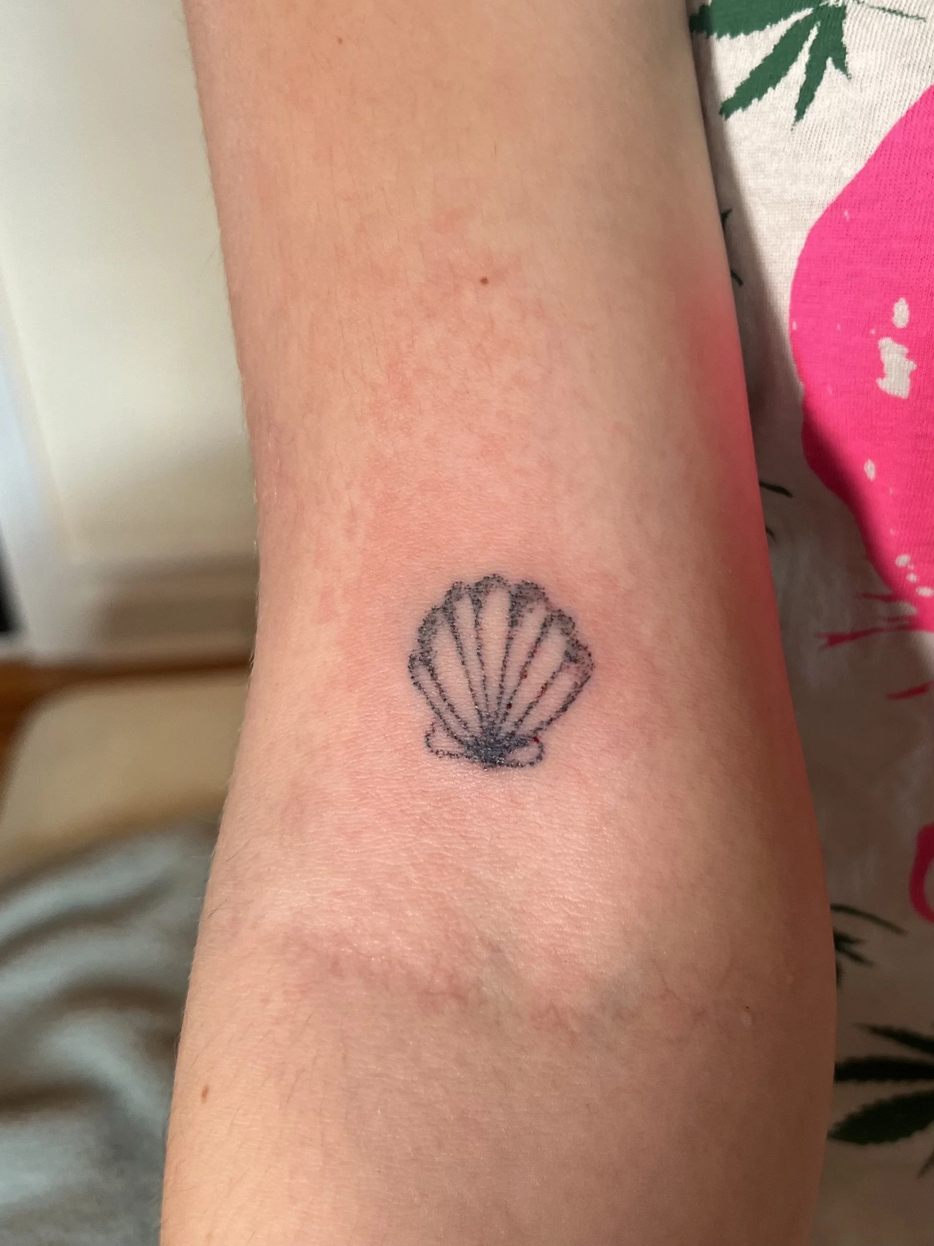 Close-up of a person's arm with a small, black tattoo of a seashell.