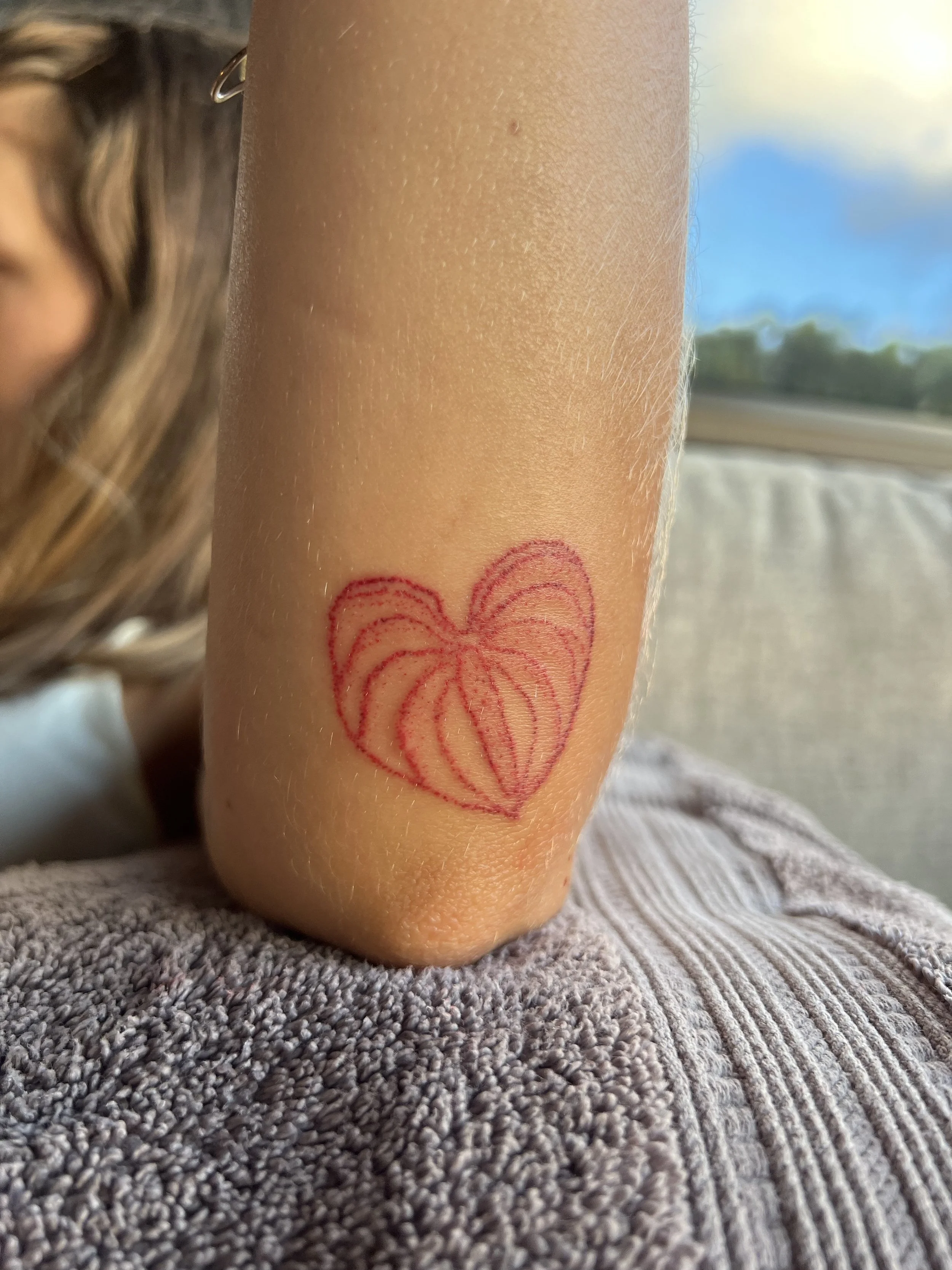 Close-up of a person's arm with a red outline tattoo of a stylized rose flower, resting on a textured gray fabric surface with a blurred outdoor background.