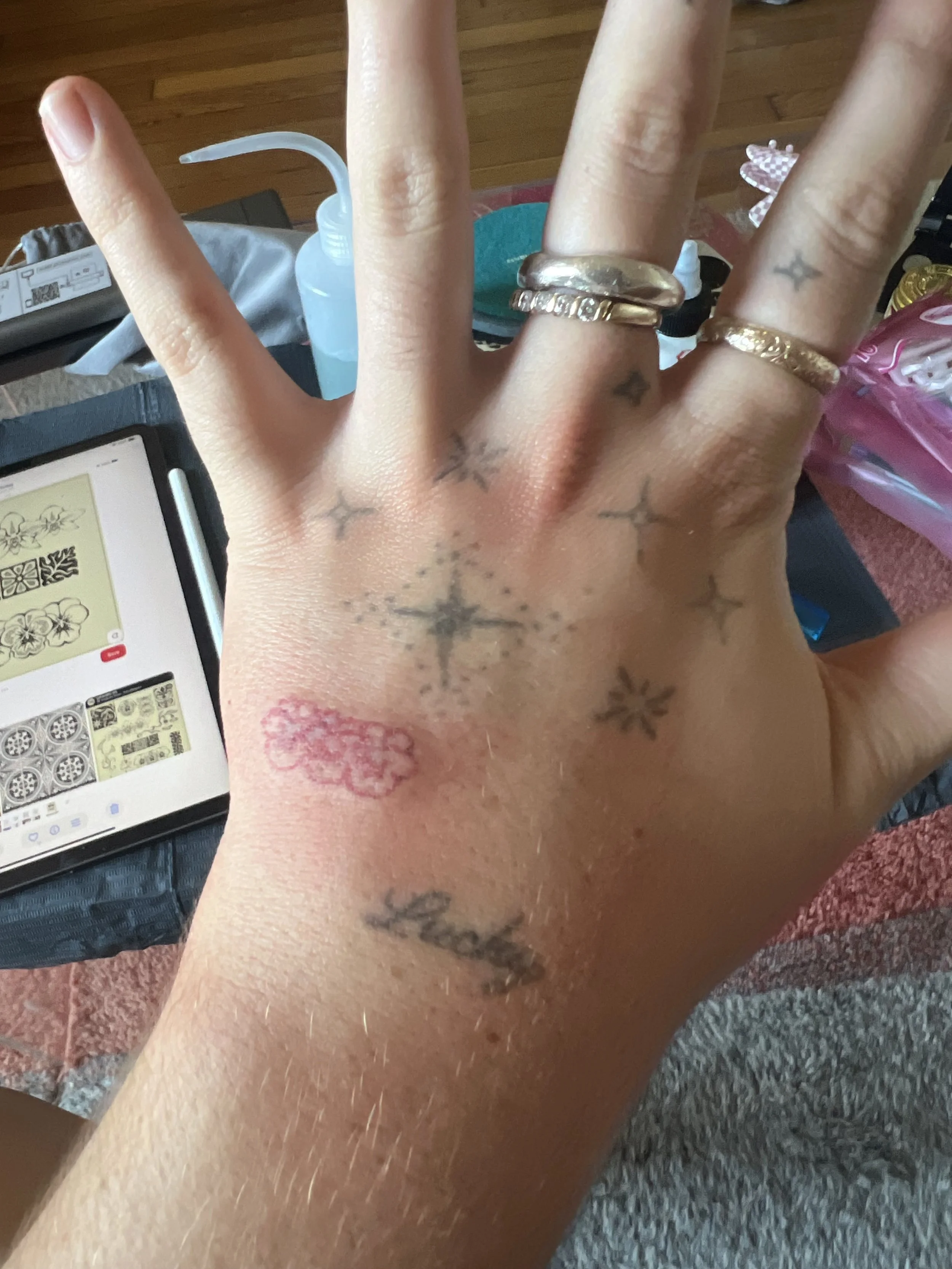 Close-up of a person's hand showing multiple rings and small star tattoos, with a pink heart tattoo on the wrist, and a smartphone displaying a design app in the background.
